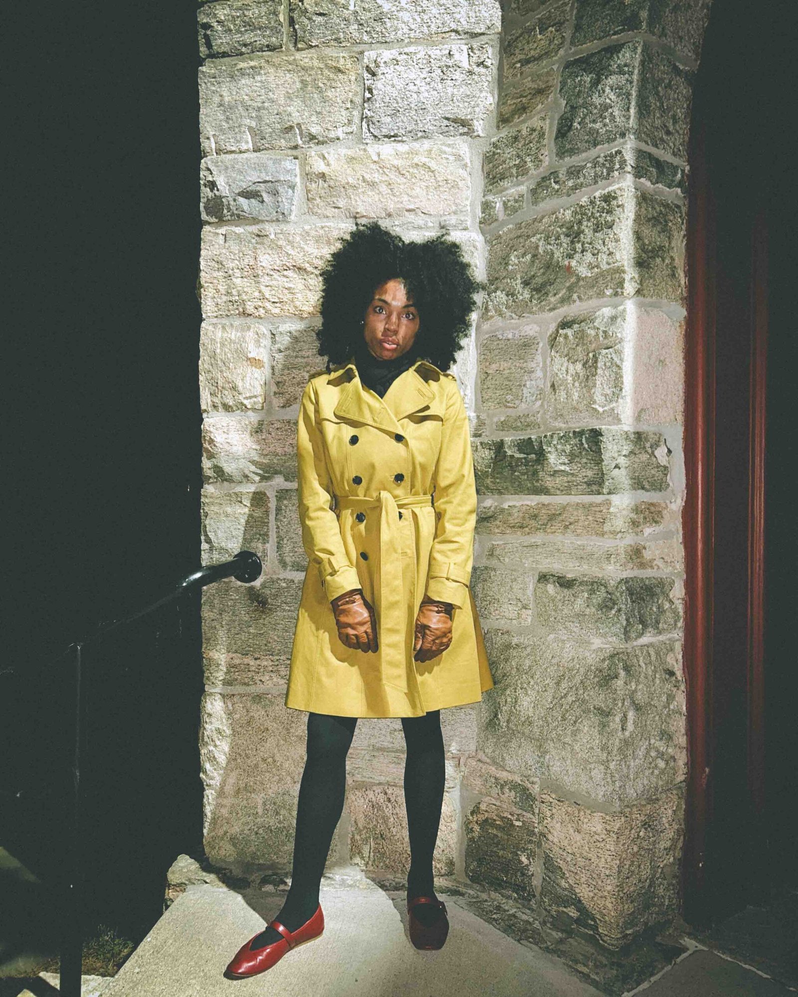 Woman wearing a yellow trench coat and red flats standing beside a stone building at night, capturing the moodiness of winter loneliness and holiday melancholy.