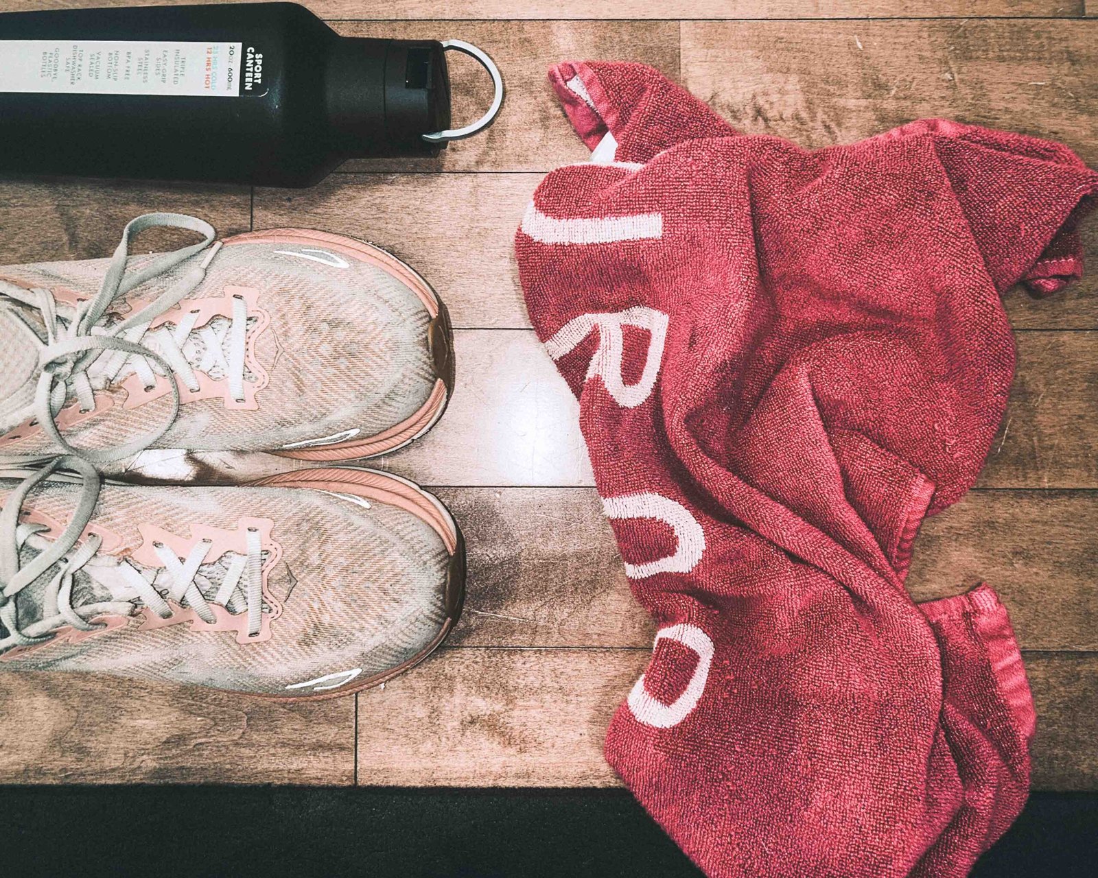 Flat lay of worn pink running shoes, a black sports water bottle, and a bright pink gym towel on a wooden floor after a workout session.