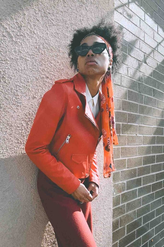 Boho fashion blogger in a red moto jacket, red knit pants, and an orange floral scarf, standing against sunlit textured walls for an editorial fall outfit photo.