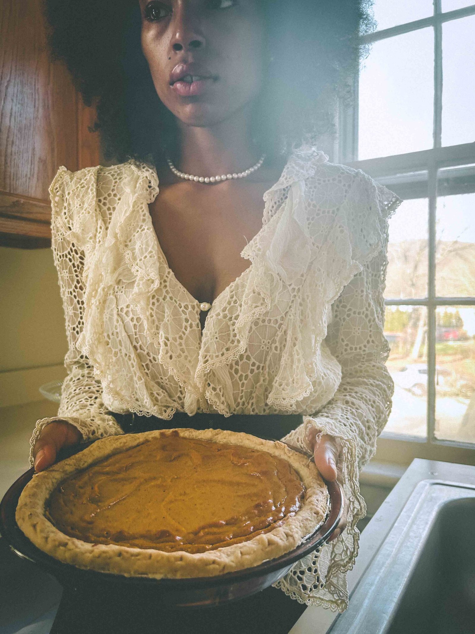 A woman in a lace blouse and pearl necklace holds a homemade sweet potato pie in soft morning light, capturing the quiet feeling of being single during the holidays.