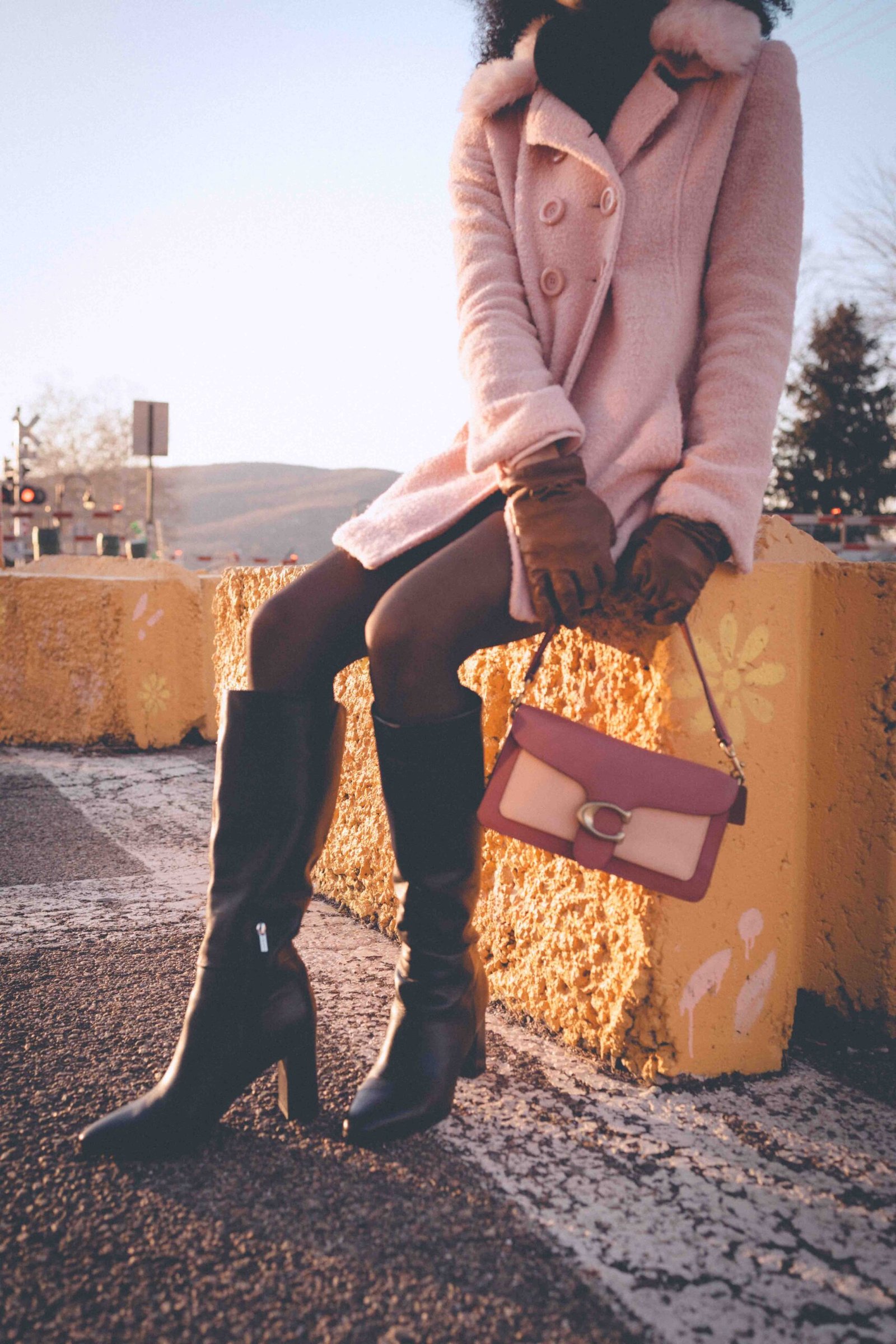 Pink wool winter coat paired with black tights, knee-high boots, leather gloves, and a blush handbag during golden hour in a city setting.