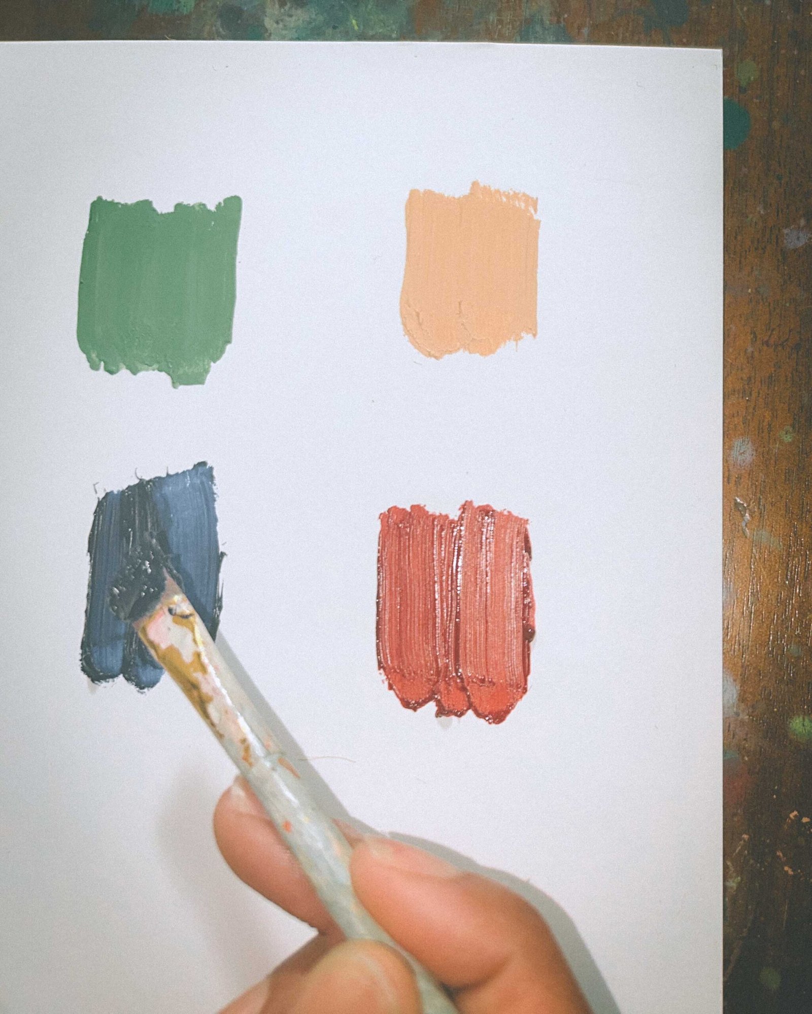 Close-up of a hand mixing navy blue oil paint on paper alongside swatches of green, peach, and brick red for an artist’s winter painting palette.
