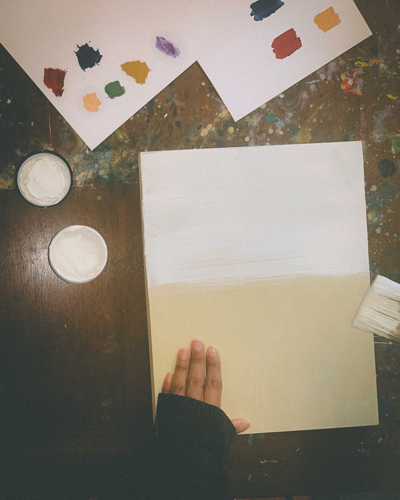 Bohemian woman artist brushing white gesso onto a wood panel on her paint-splattered table while building the foundation for a winter art series.