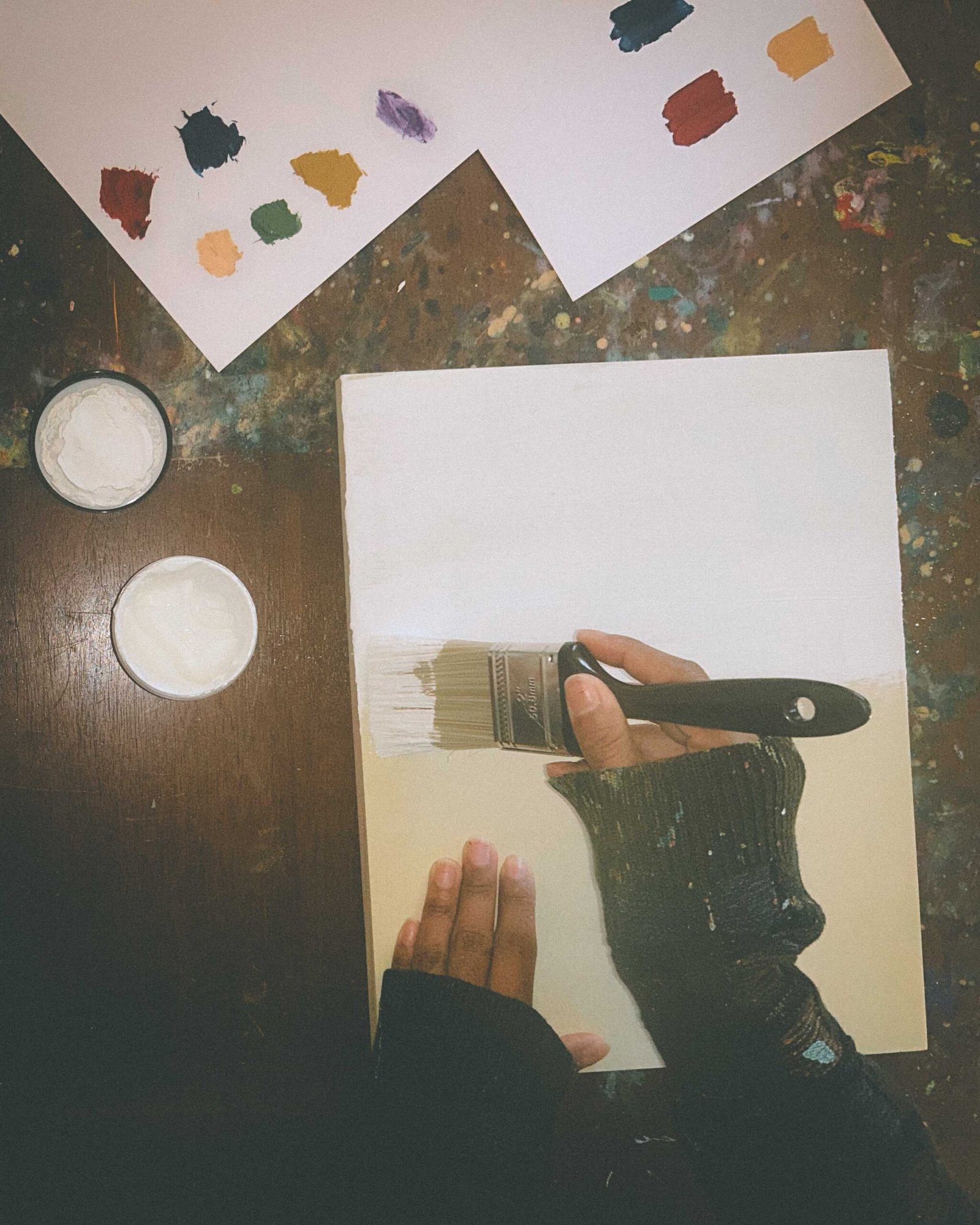 Artist’s hand resting on an unprimed wood panel beside matte medium and paint swatches as part of preparing surfaces for a new mixed-media art series.