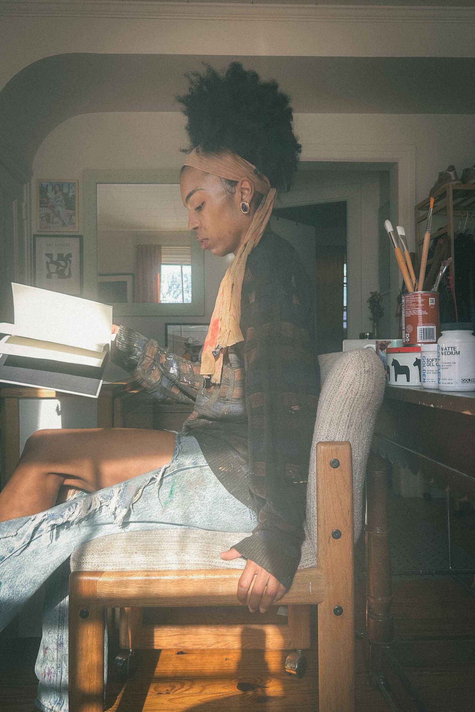 Artist, healing through art, sitting in her winter art studio reading “The Creative Act” with afternoon sunlight on her face, wearing paint-splattered jeans and a cozy scarf.