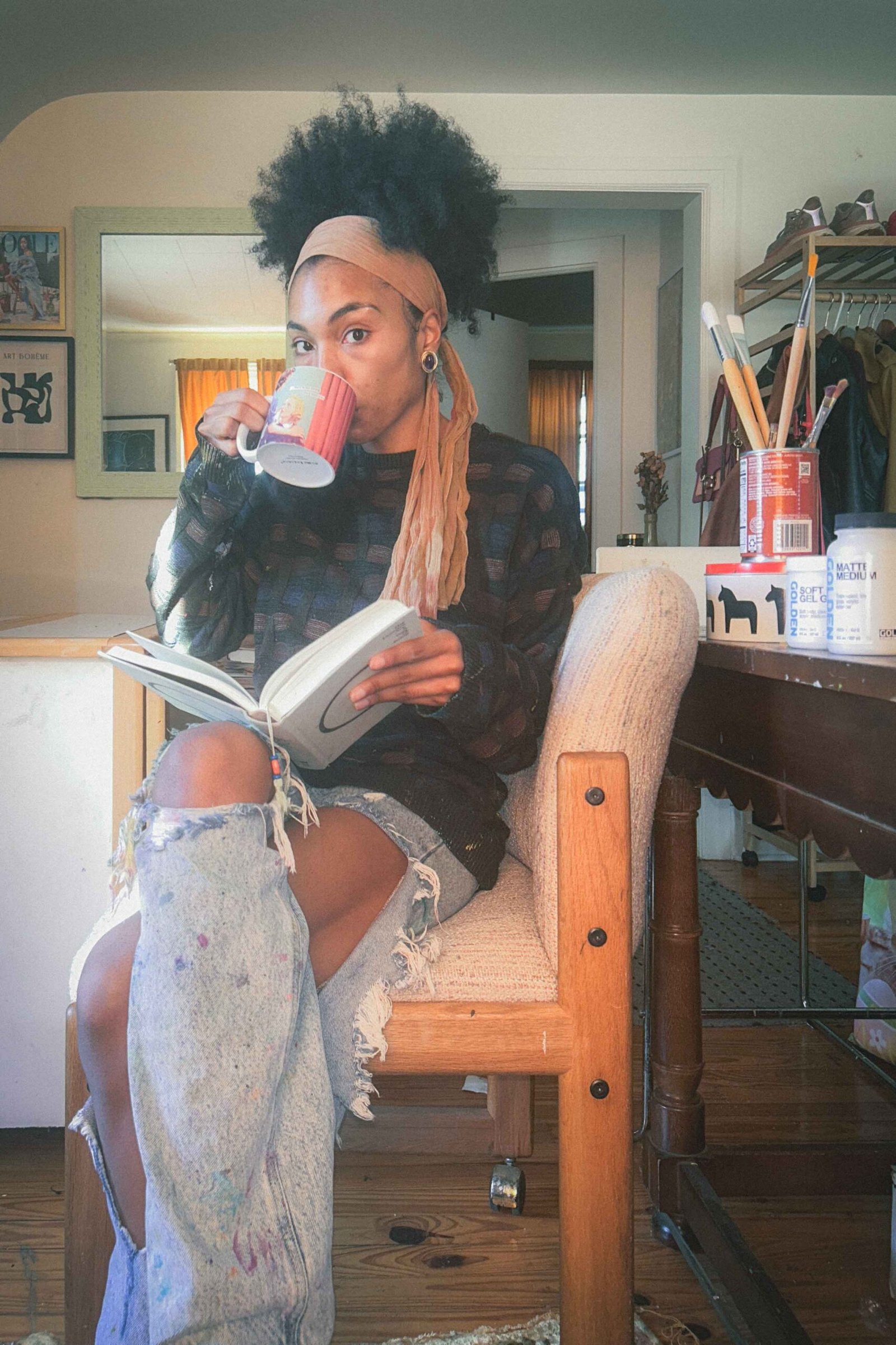 Bohemian woman artist in her winter art studio sipping coffee and reading a book in paint-splattered jeans, surrounded by her brushes and painting supplies.