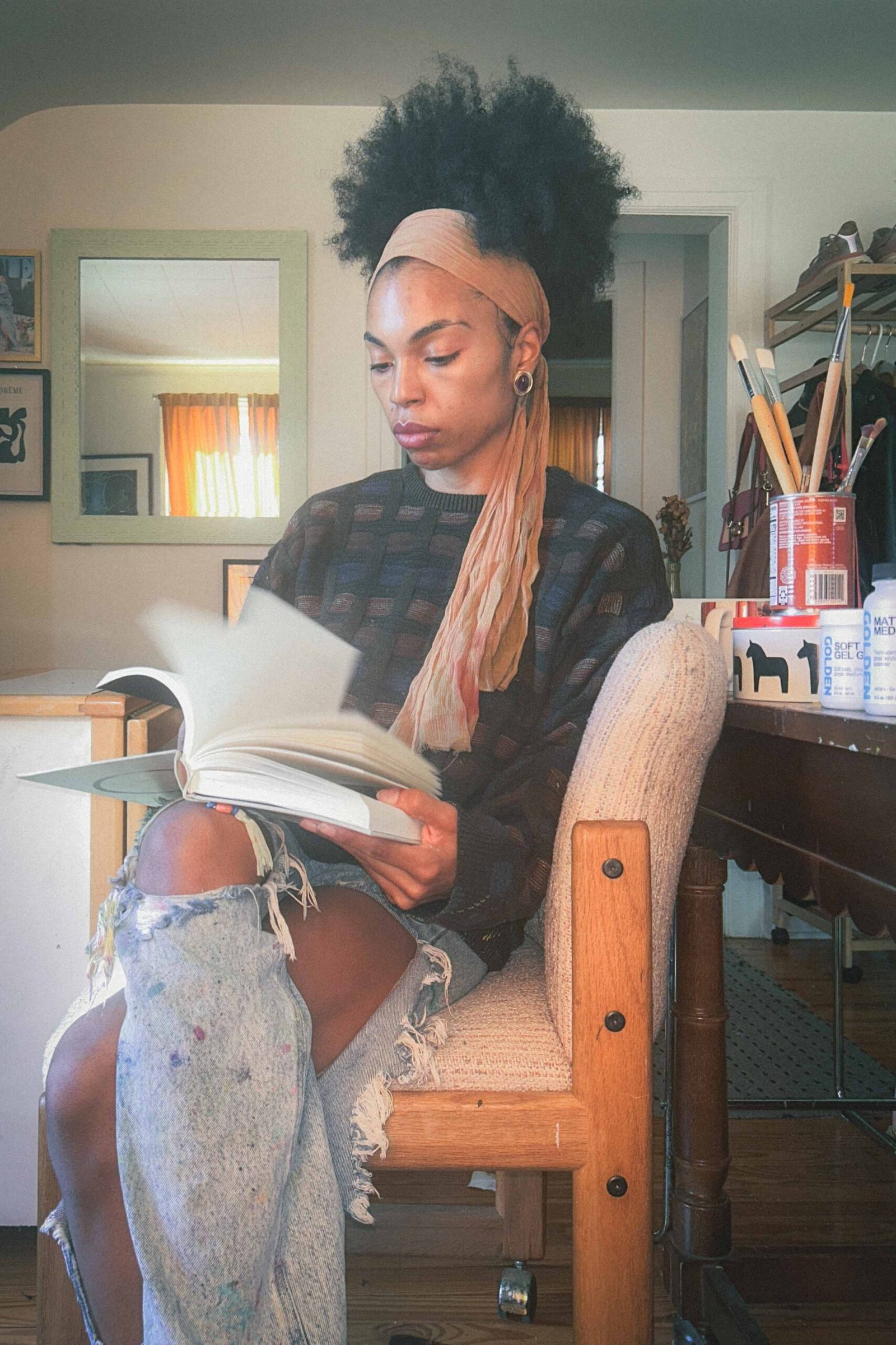 Artist reading “The Creative Act: A Way of Being” in her studio, pages in motion, framed by warm winter light and studio tools.