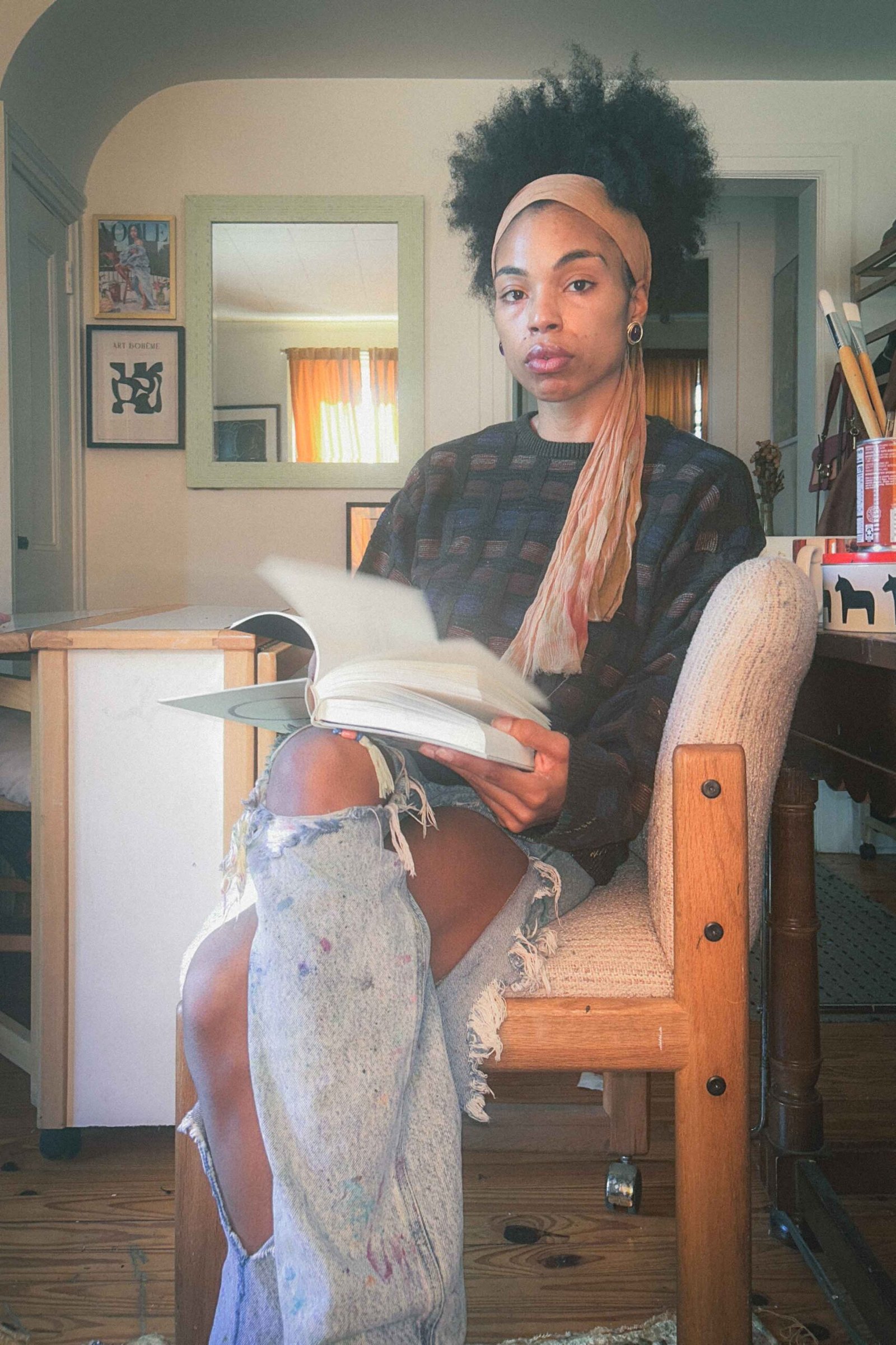 Artist sitting in her winter studio, holding an open book with a calm direct gaze, wearing distressed paint-stained jeans and an oversized sweater.