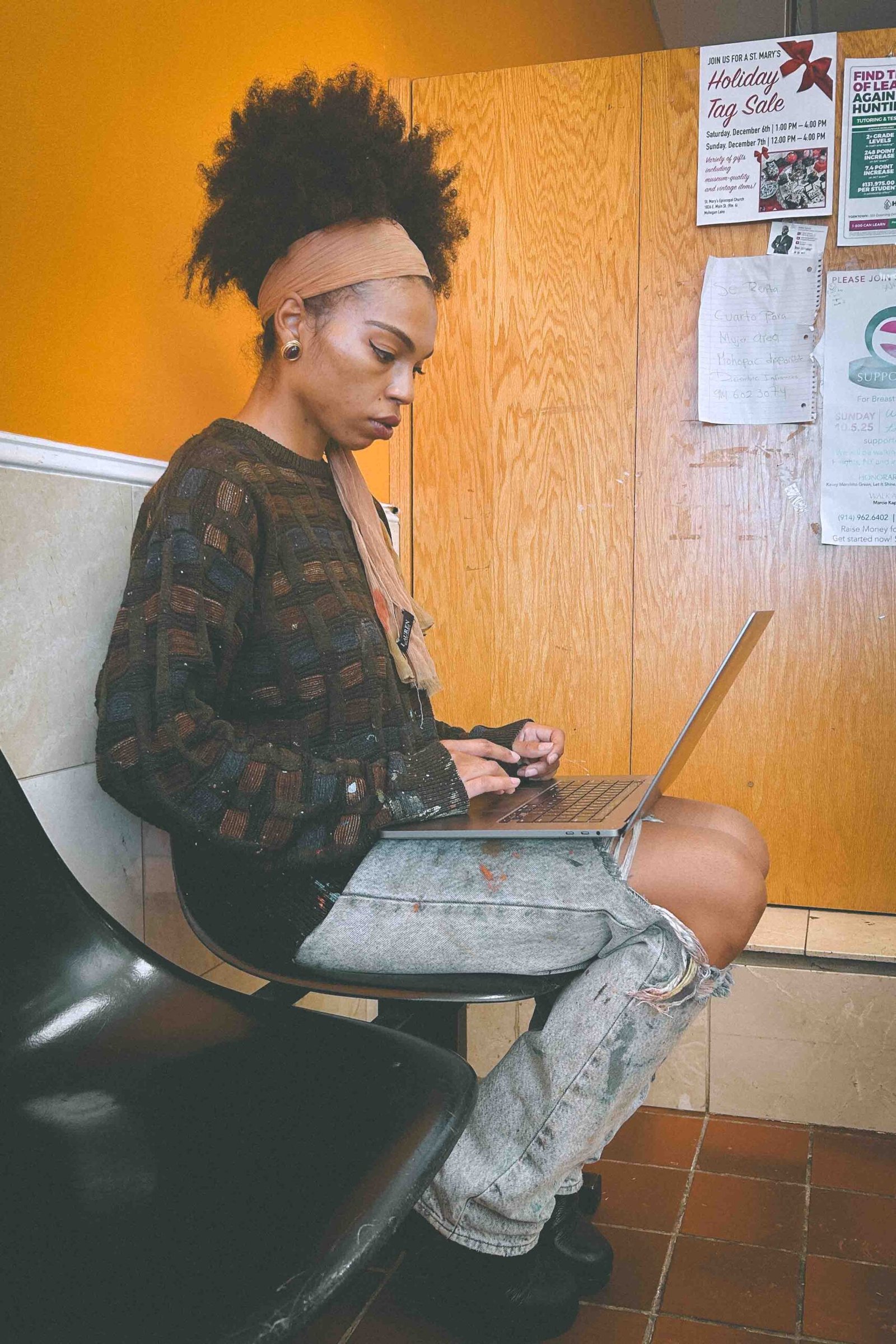 Bohemian woman artist working on her laptop at a laundromat in Hudson Valley, New York wearing her paint-splattered jeans and knit sweater while balancing real life and art-making.