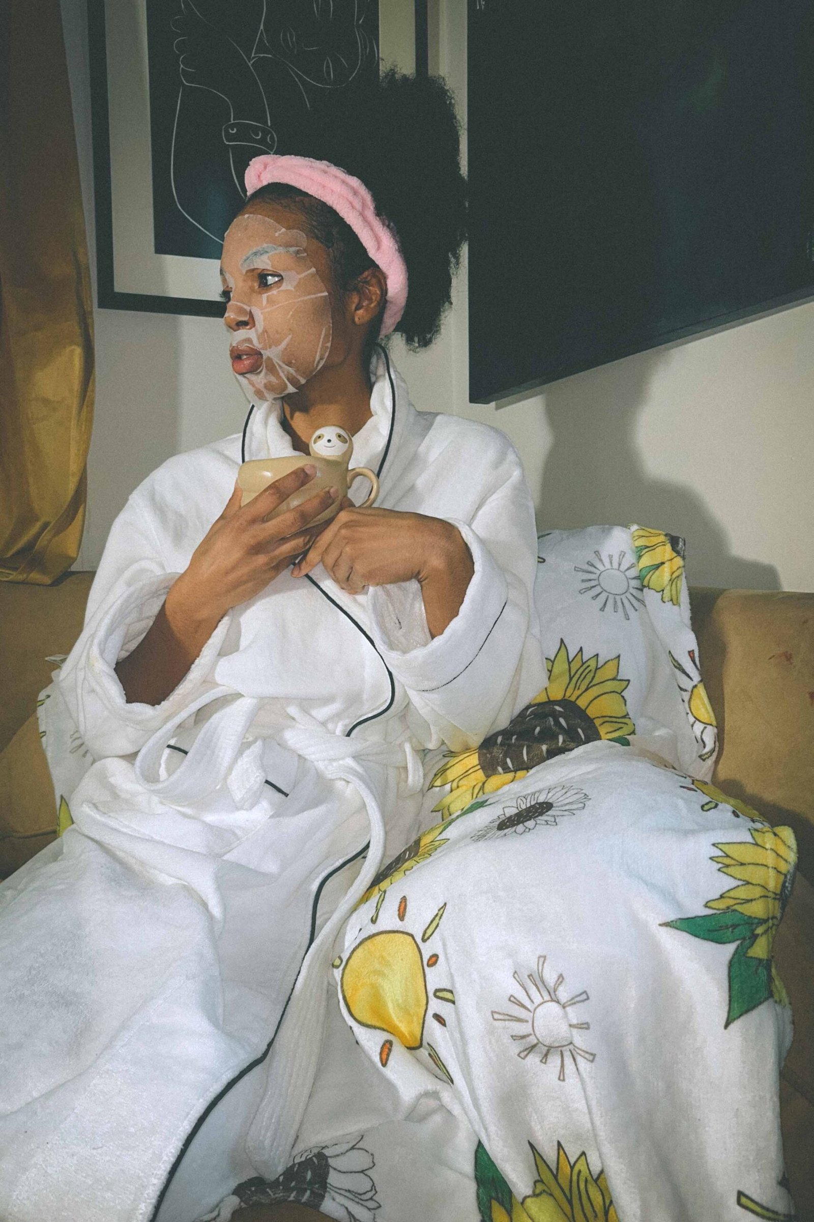 A bohemian woman seated on a couch in a cozy robe, holding a mug and wearing a sheet mask as part of her daily morning hydration routine.