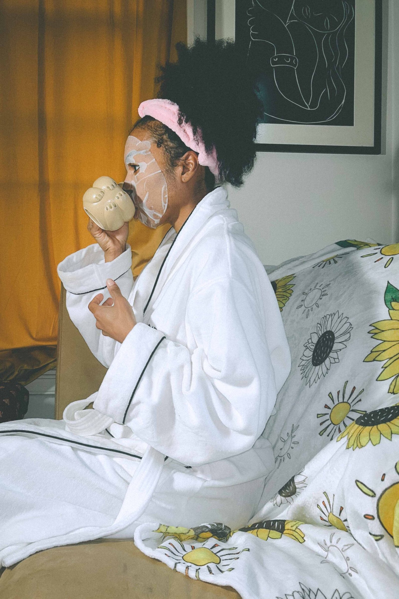A bohemian woman in a white robe and pink headband drinking coffee while wearing a hydrating sheet mask during her slow morning skincare ritual.