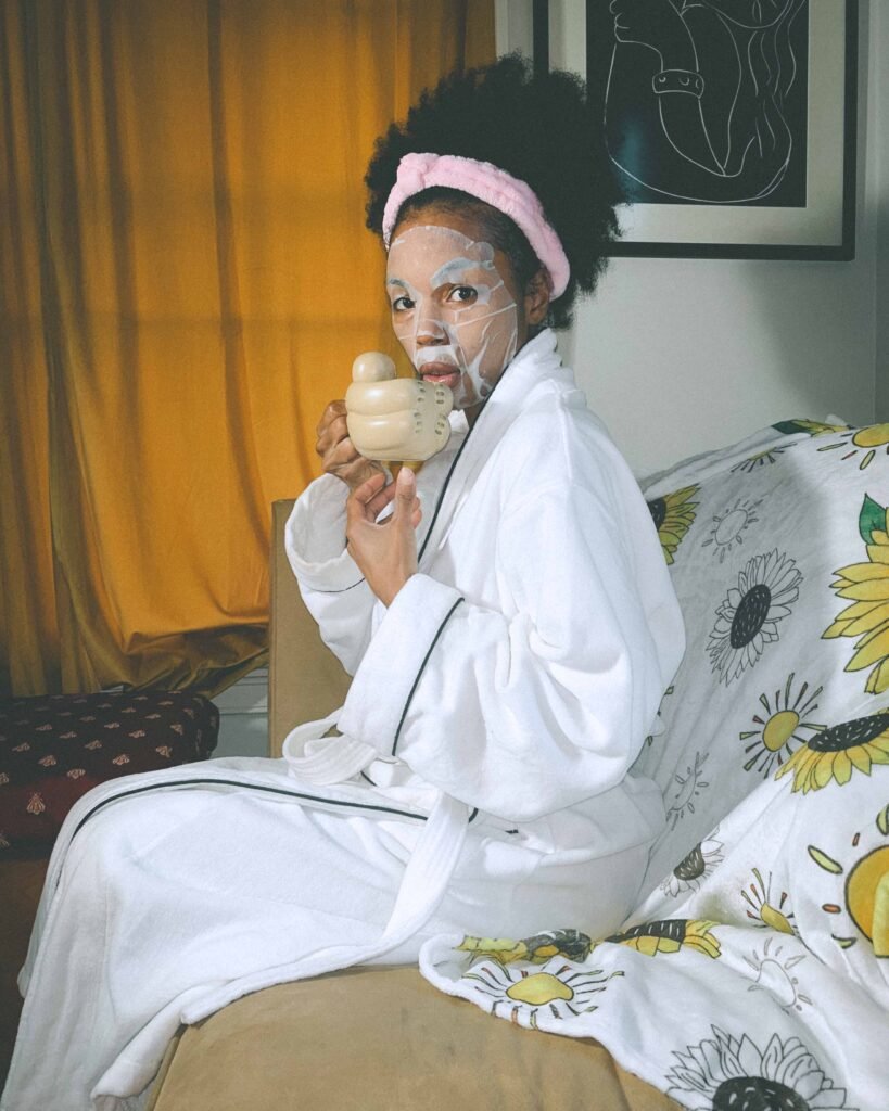 Bohemian woman wearing a white robe and pink headband sipping coffee while sitting on a couch with a sheet mask on, during her cozy morning skincare routine.
