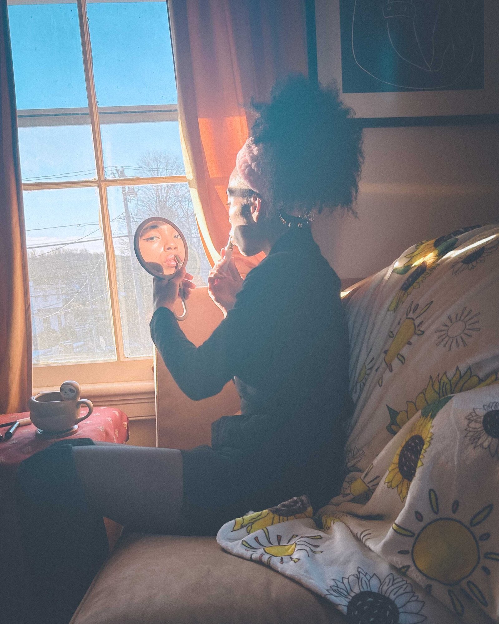 A bohemian woman applying makeup by a sunlit window, holding a small mirror that reflects her face as she begins her simple morning makeup routine.
