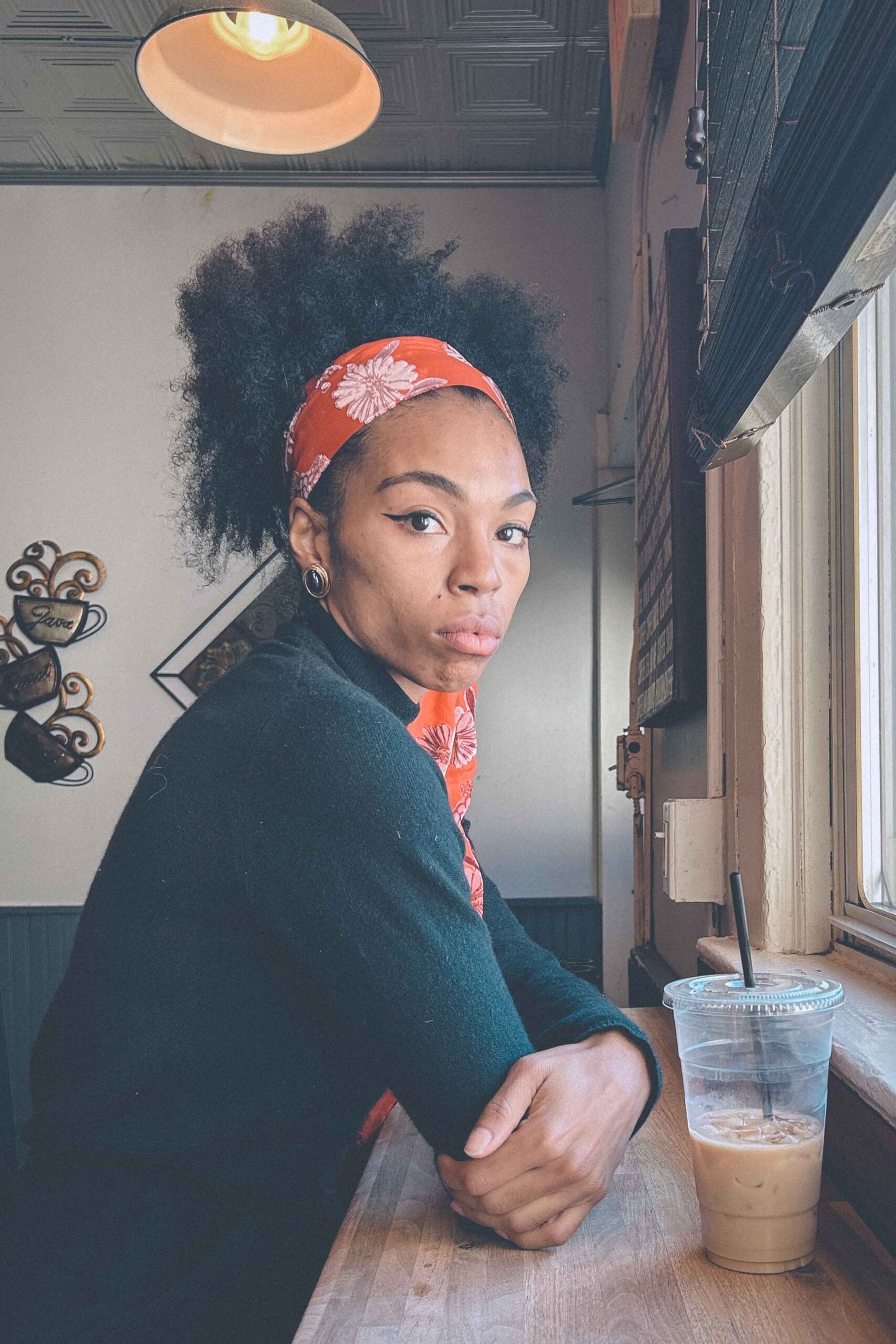 Bohemian woman sitting at a café counter by a window, wearing a floral headscarf and black sweater, with an iced coffee beside her, looking toward the camera.