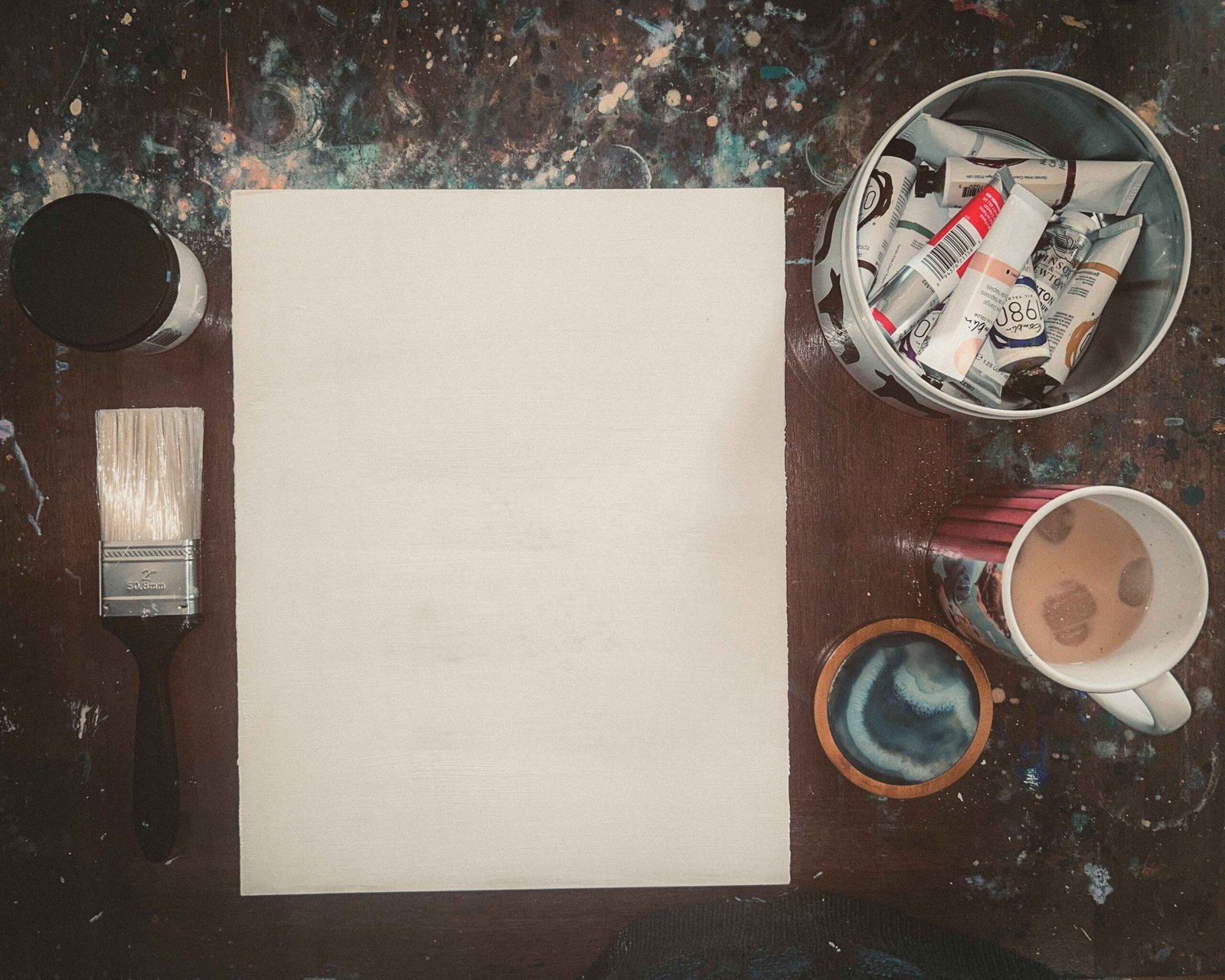 Flatlay of a primed wood panel surrounded by Gamblin oil paints, a large brush, and a mug of iced coffee on a paint-splattered studio table.