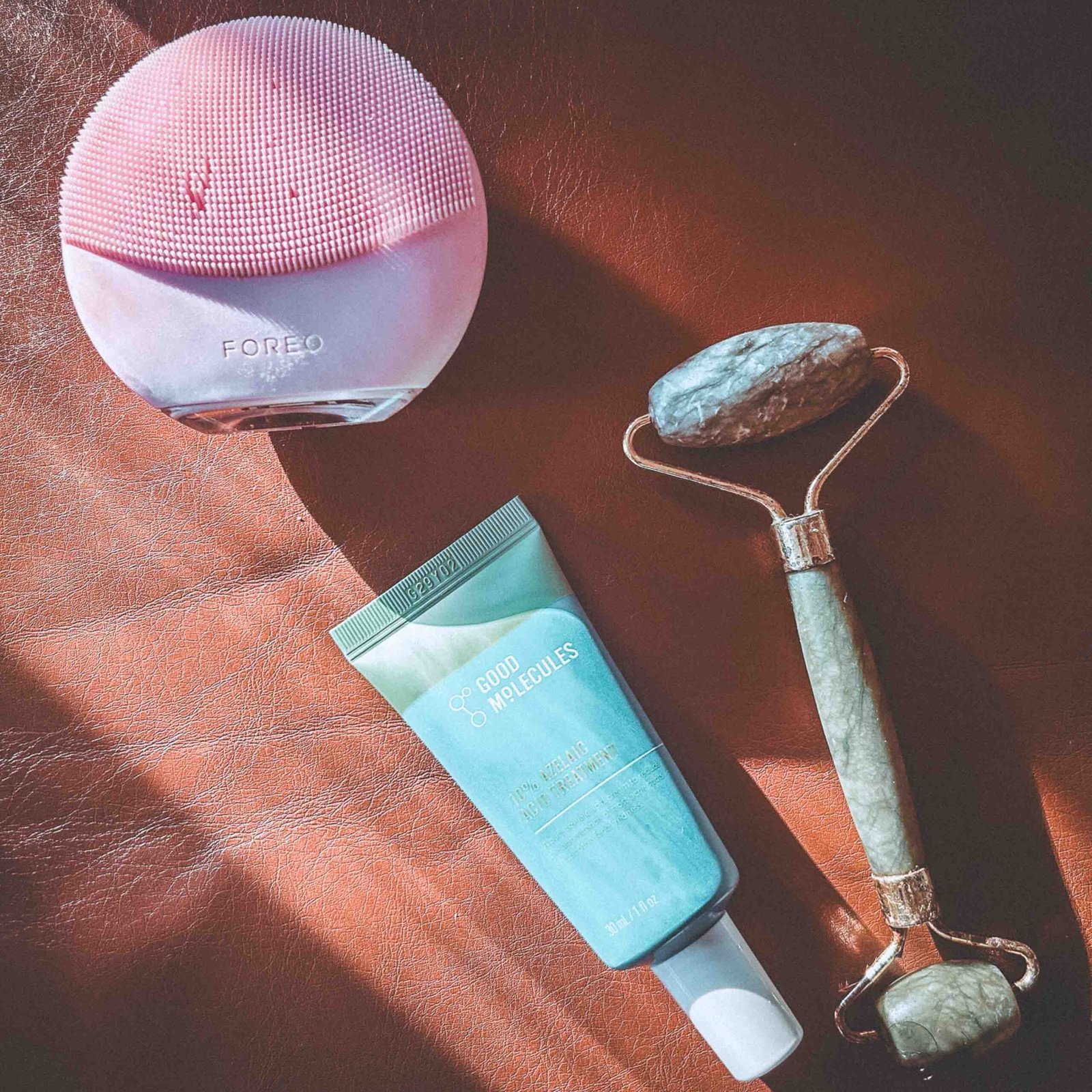 Flatlay of a pink Foreo cleansing device, Good Molecules azelaic acid treatment, and jade facial roller displayed on a brown leather surface in soft morning light.