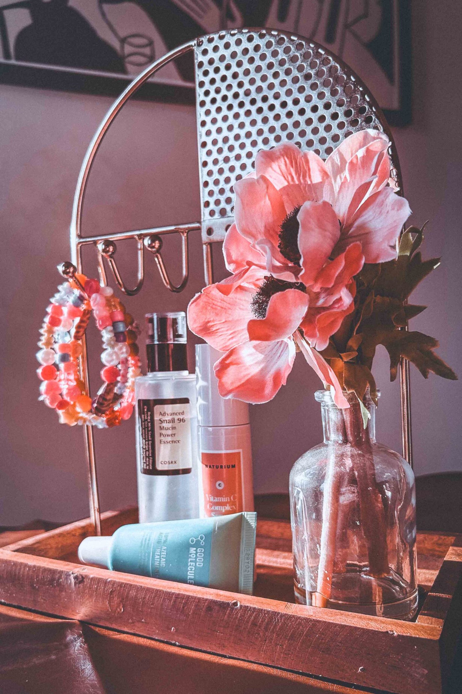 Bohemian display of morning skincare routine essentials featuring COSRX Snail Mucin Essence, Naturium Vitamin C Serum, Good Molecules Azelaic Acid treatment, a pink faux flower in a glass vase, and beaded bracelets arranged on a wooden tray.