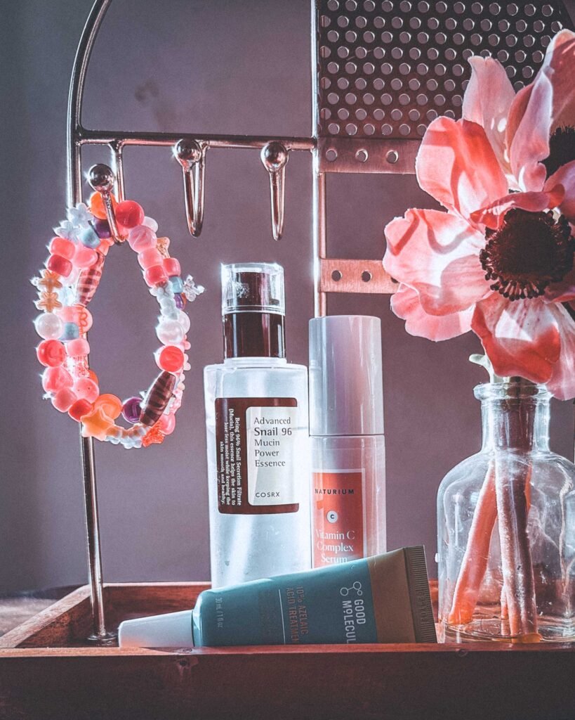 Close-up bohemian skincare flatlay with COSRX Snail Mucin Essence, Naturium Vitamin C Serum, Good Molecules Azelaic Acid treatment, beaded bracelets, and a pink faux flower on a wooden tray in soft natural light.
