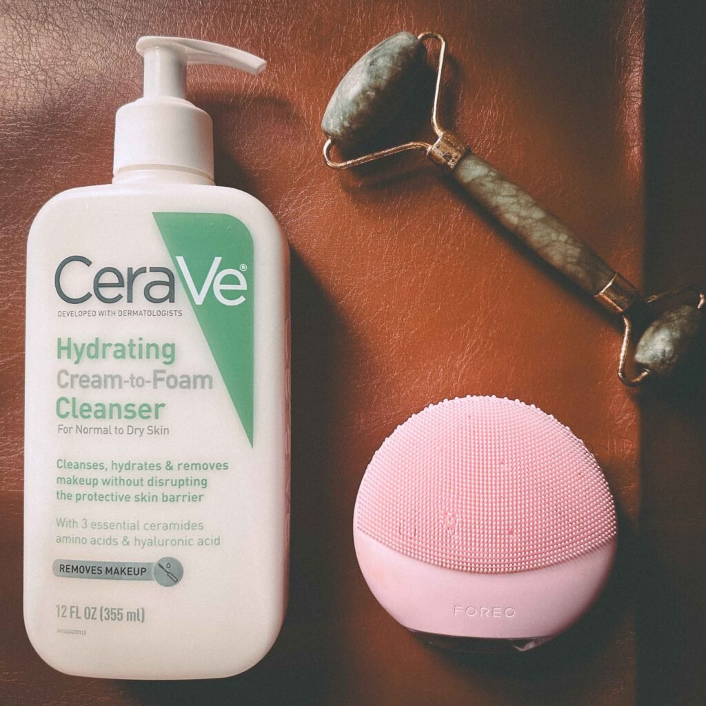 Flatlay of CeraVe Hydrating Cream-to-Foam Cleanser, a pink Foreo cleansing device, and a jade facial roller arranged on a brown leather surface in soft natural light.