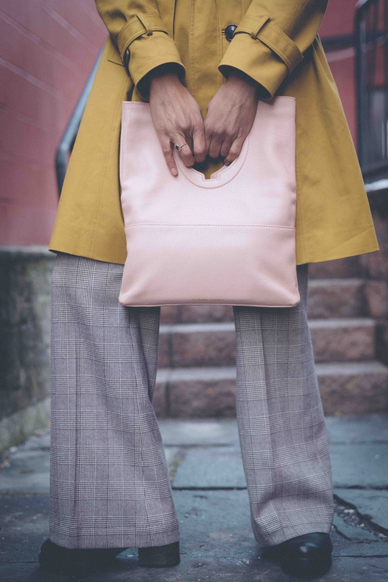 Soft pink leather handbag held at waist level, paired with a mustard trench coat and gray plaid trousers, styled for a polished boho-luxe winter outfit.