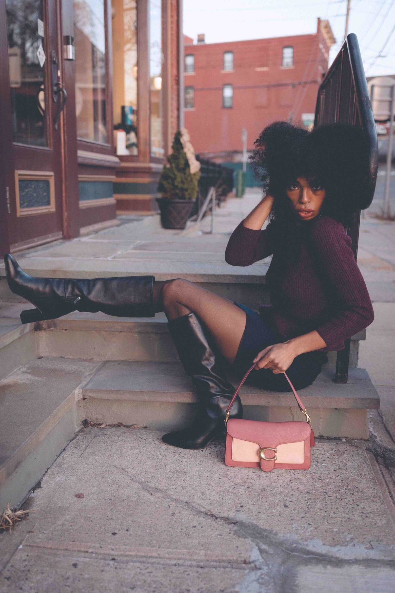 Burgundy knit sweater styled with a black mini skirt, sheer tights, knee-high leather boots, and a pink shoulder bag on city steps in winter.