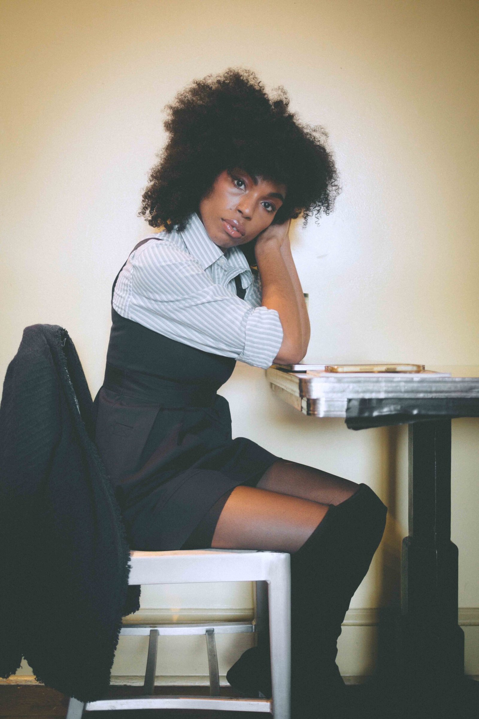 Art girl winter outfit featuring a thrifted navy pinafore dress layered over a striped button-down, worn with over-the-knee boots and a vintage chain bag on a quiet winter street.