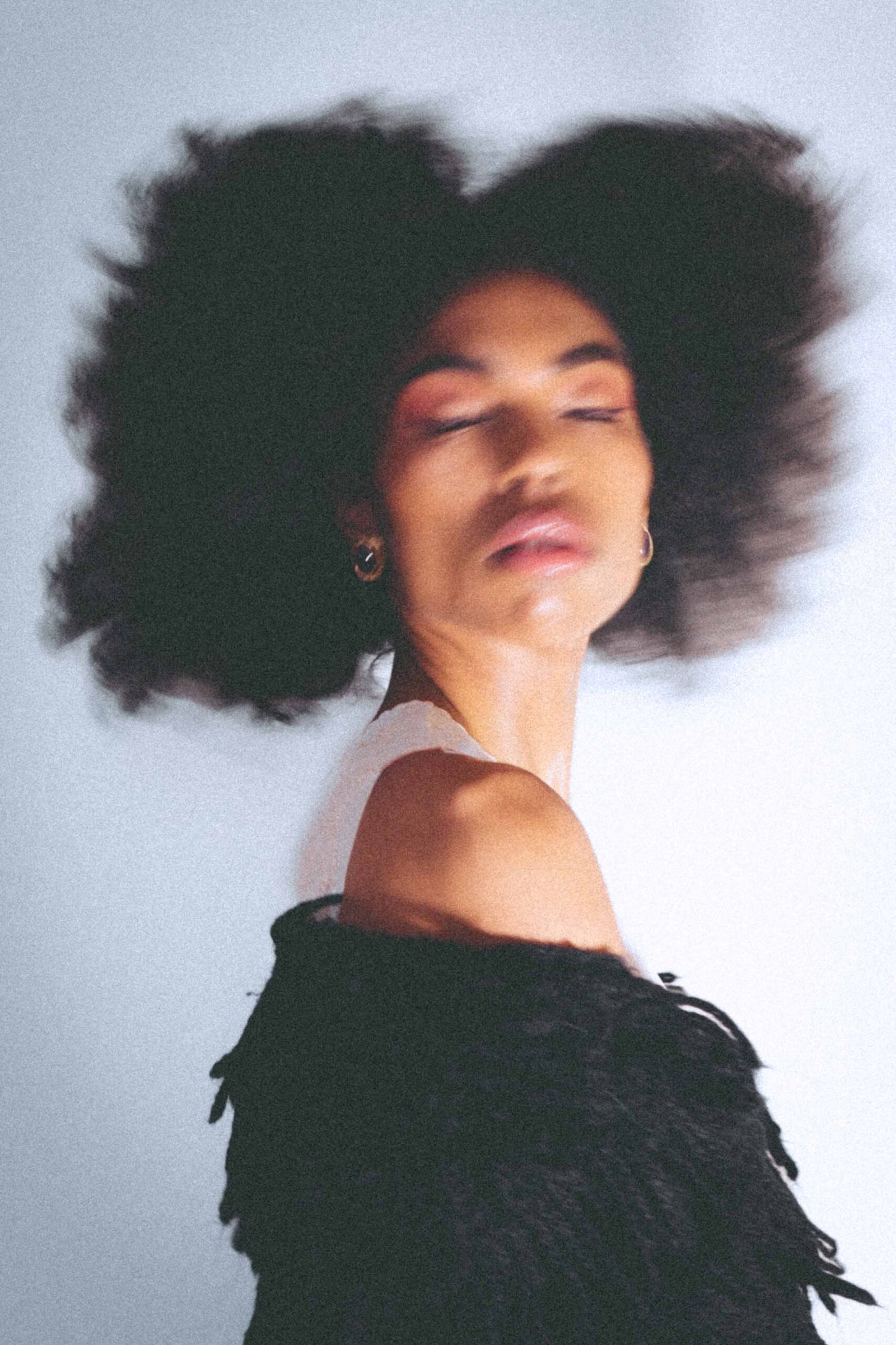 Soft motion-blur portrait of a woman with natural hair and minimal makeup, eyes closed, expressing movement and becoming through an editorial art girl winter self-portrait.