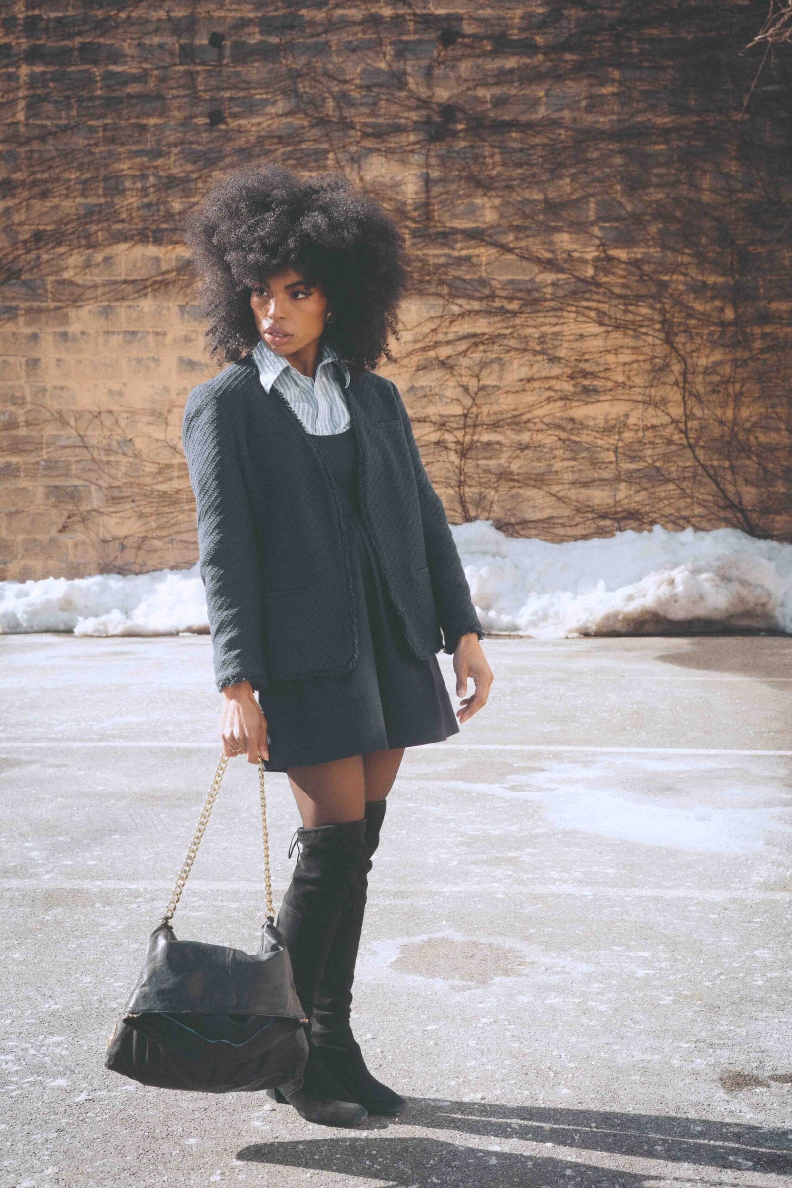 Art girl winter outfit featuring a thrifted navy pinafore dress layered over a striped button-down, worn with over-the-knee boots and a vintage chain bag on a quiet winter street.