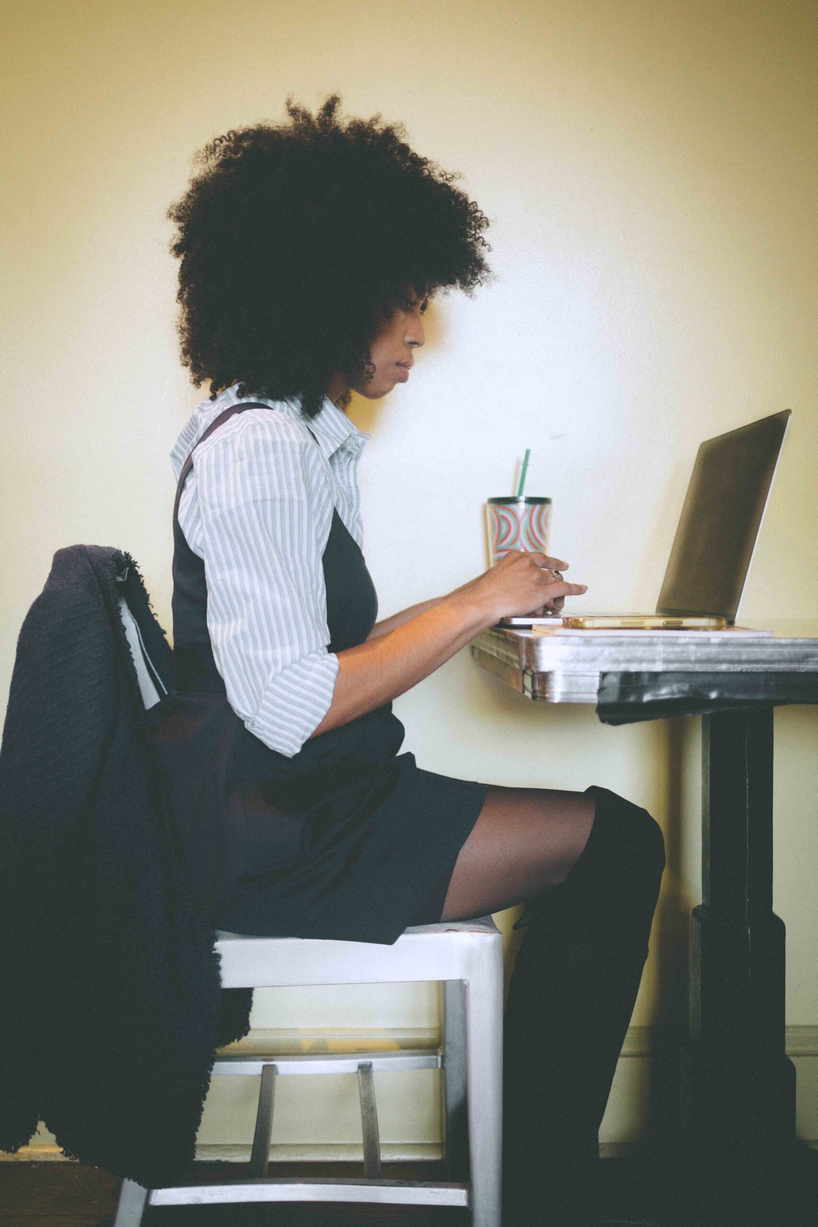 Creative woman in a thrifted pinafore dress layered over a striped button-down, sitting at a café-style table and working on a laptop during winter, embodying the art girl winter aesthetic.