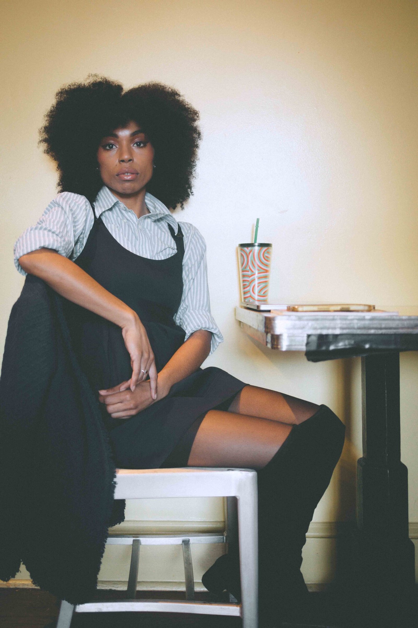 Art girl winter outfit styled indoors, featuring a thrifted black pinafore layered over a striped button-down, knee-high boots, and a quiet café-inspired setting.