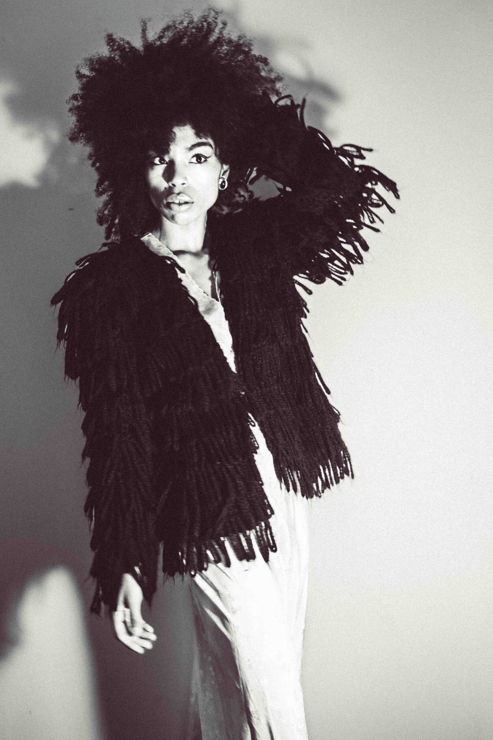 Black and white editorial self-portrait of a woman wearing a textured fringe jacket, arm lifted, capturing movement and tension within the art girl winter aesthetic.