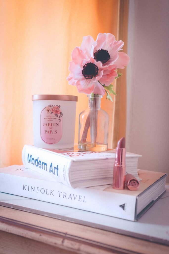 Still life of books, lipstick, flowers, and a candle, representing little luxuries and creative living at home.