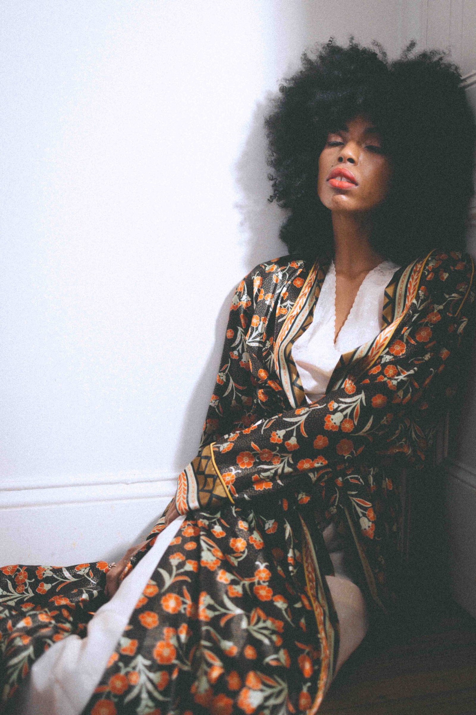 Woman in a vintage floral robe and soft makeup, leaning against a wall indoors, representing private glamour and the quiet intimacy of art girl winter style.