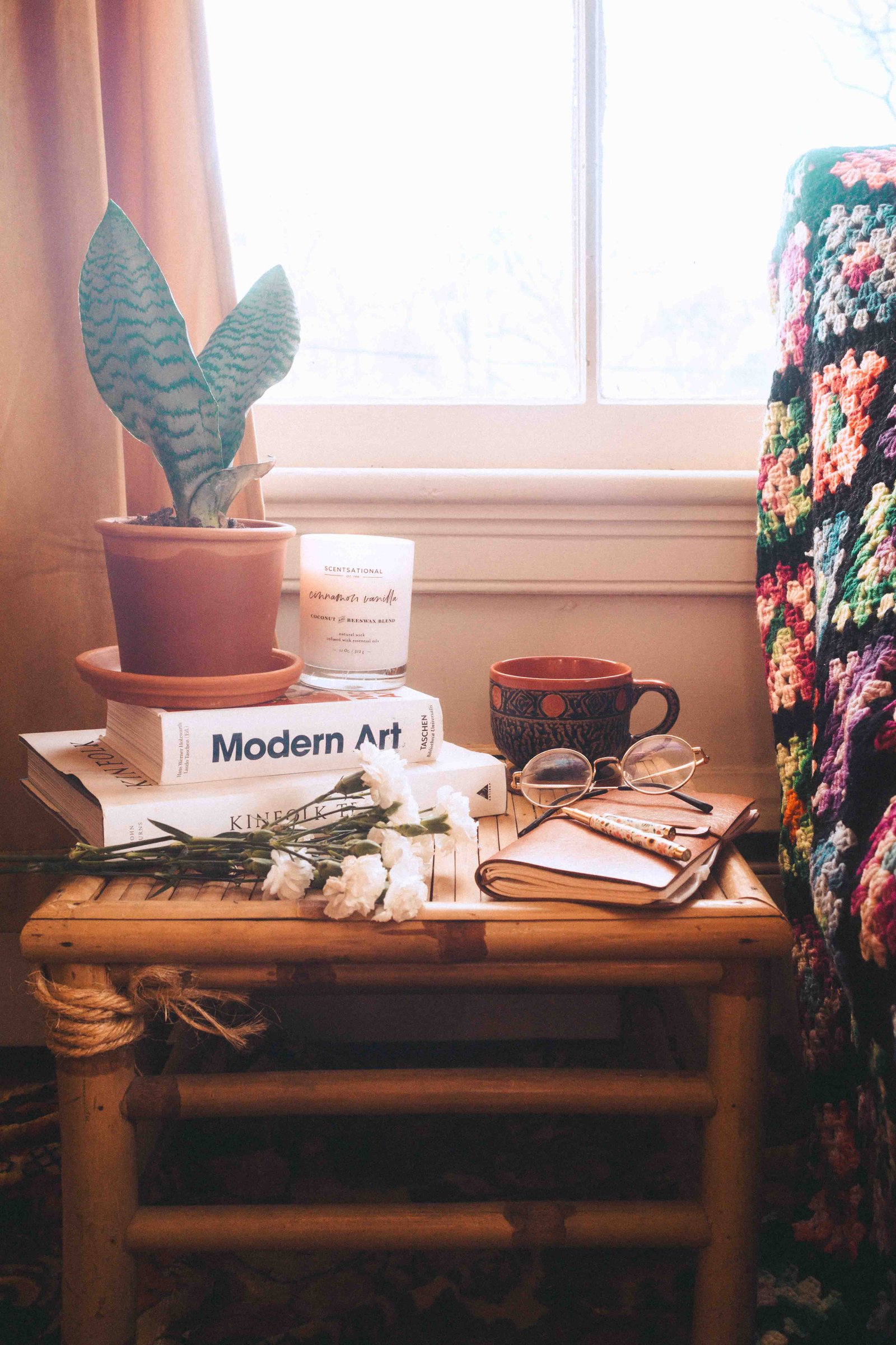 Bohemian apartment side table with books, plant, candle, and crochet blanket