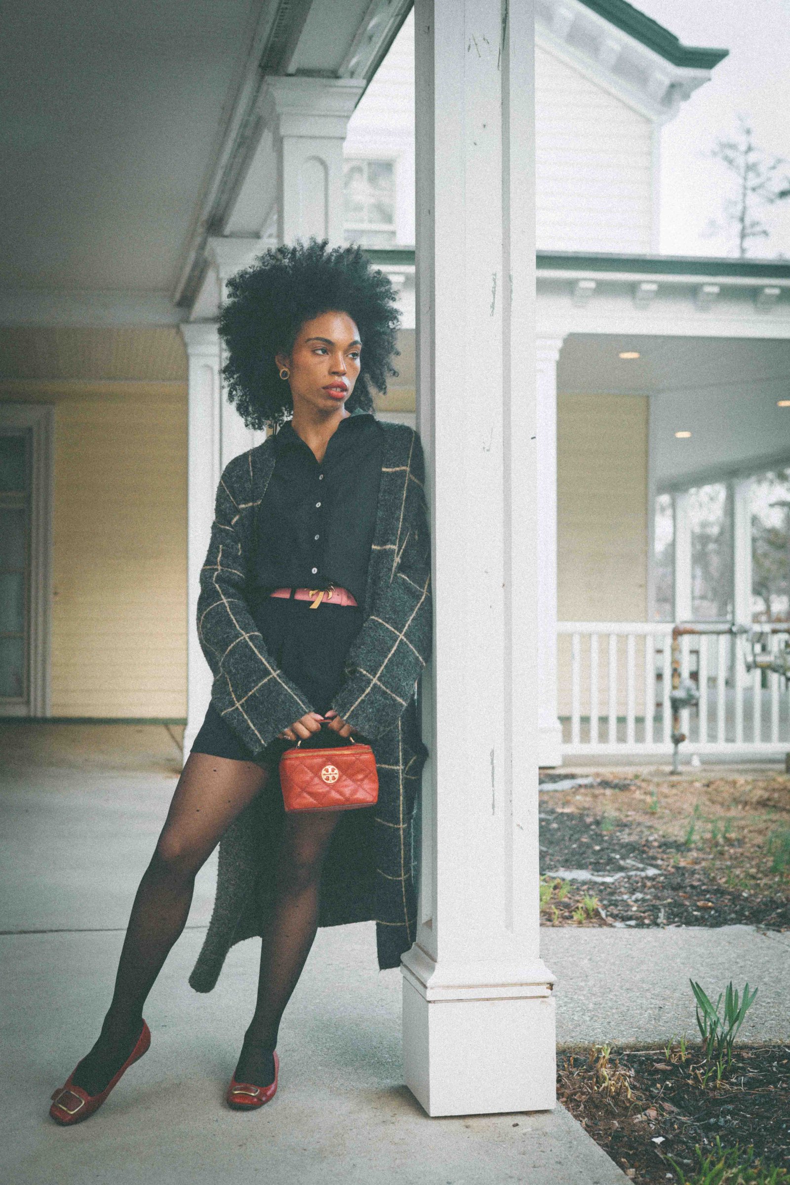 Black woman leaning on Victorian house porch column in plaid cardigan and sheer tights, bohemian bungalow style