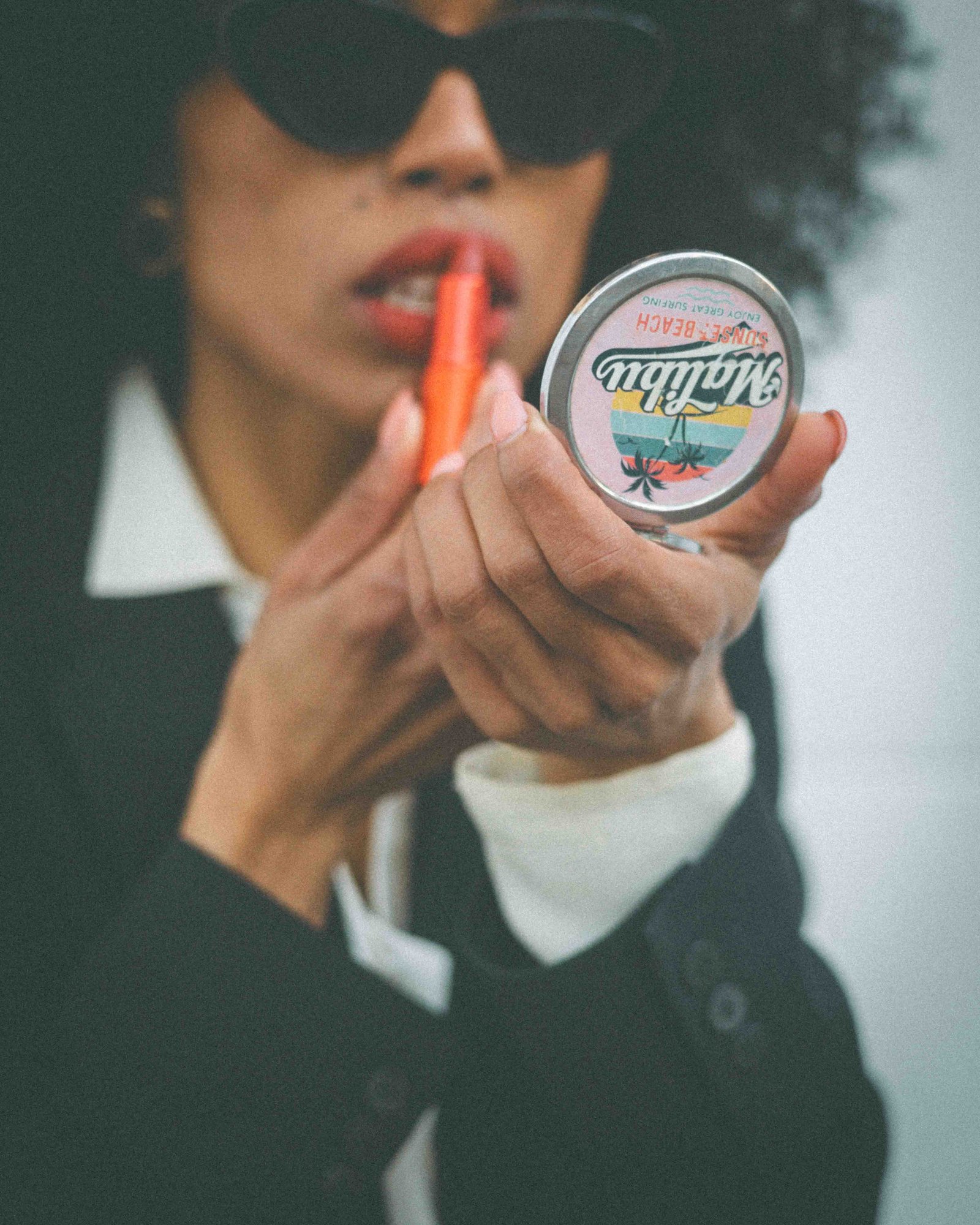 Black woman holding vintage Malibu compact mirror while applying lipstick, dressing with intention, editorial detail