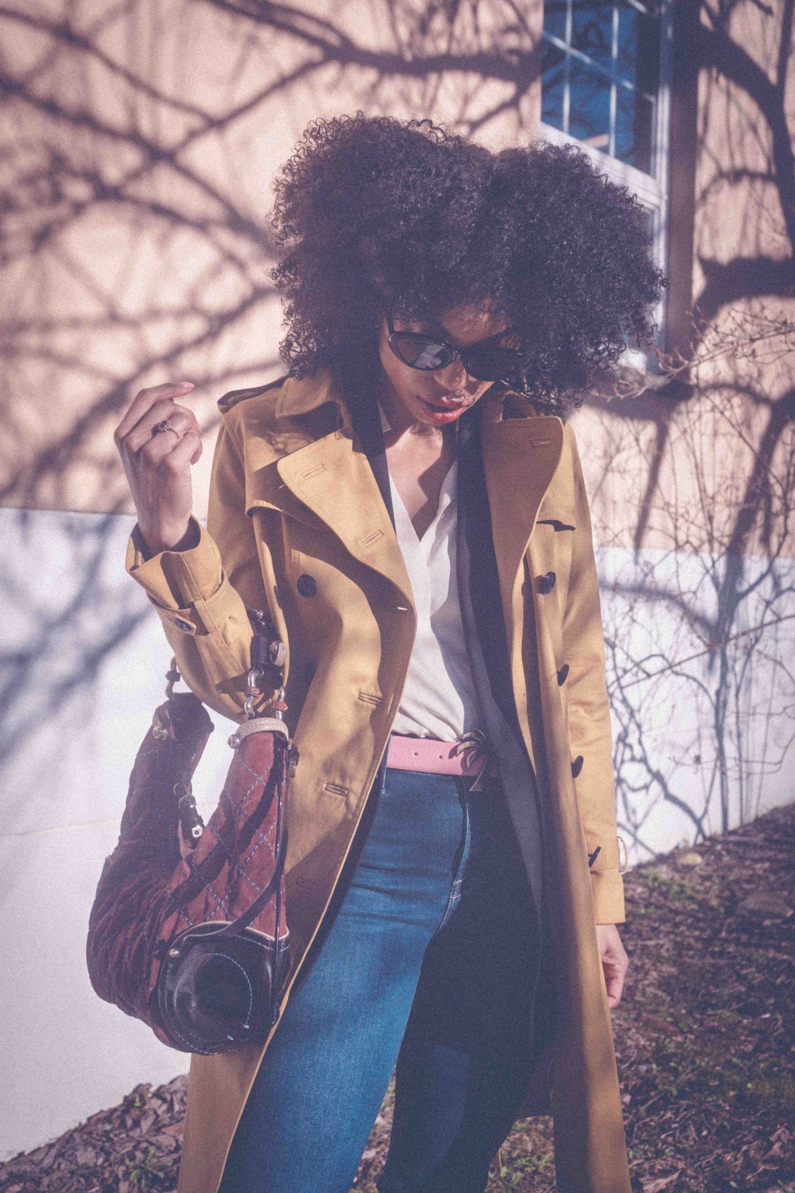Black woman in camel trench coat looking down, natural hair, moody editorial style portrait.
