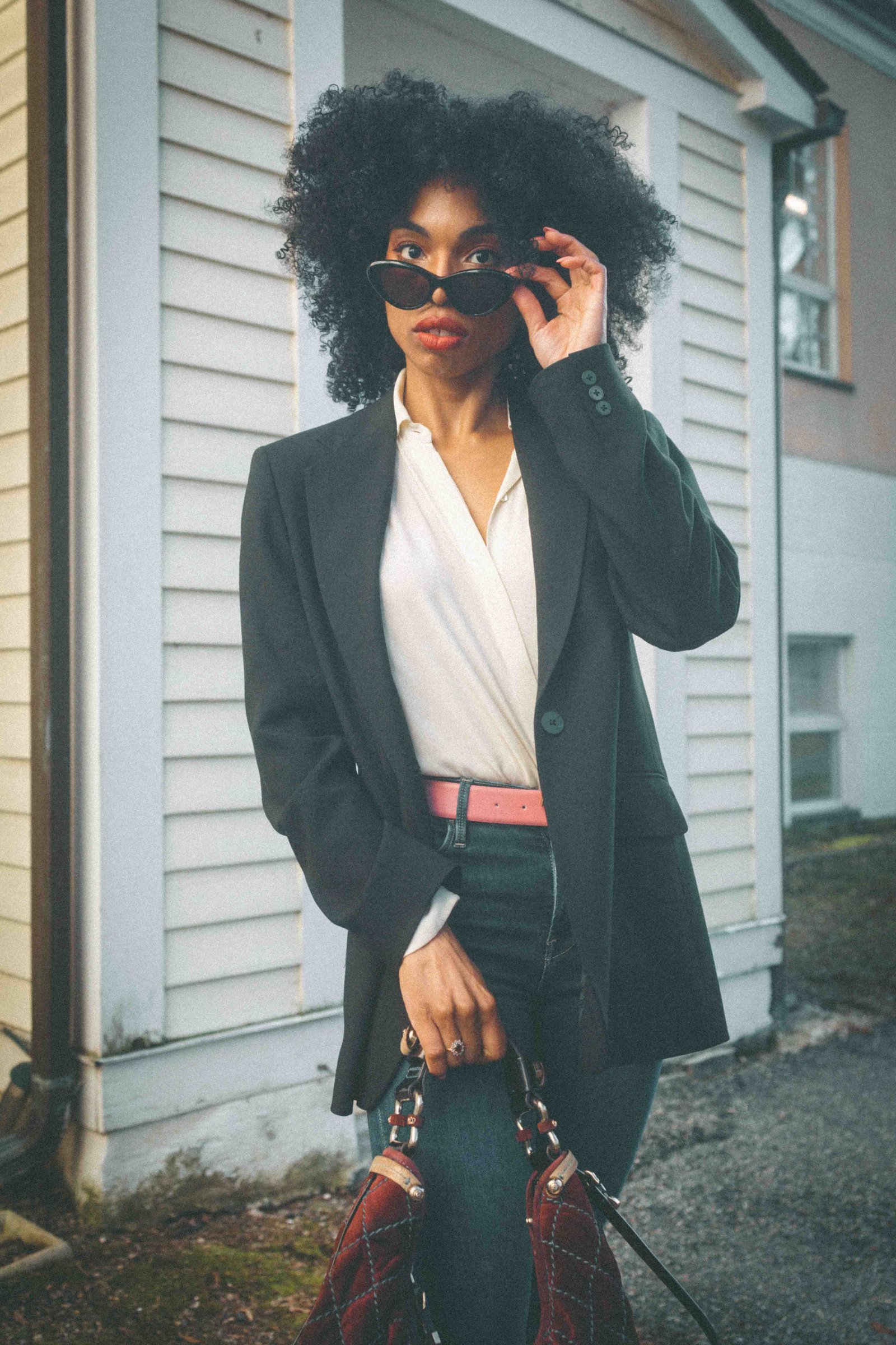 Black woman in dark blazer peering over cat eye sunglasses, editorial fashion portrait, dressing with intention