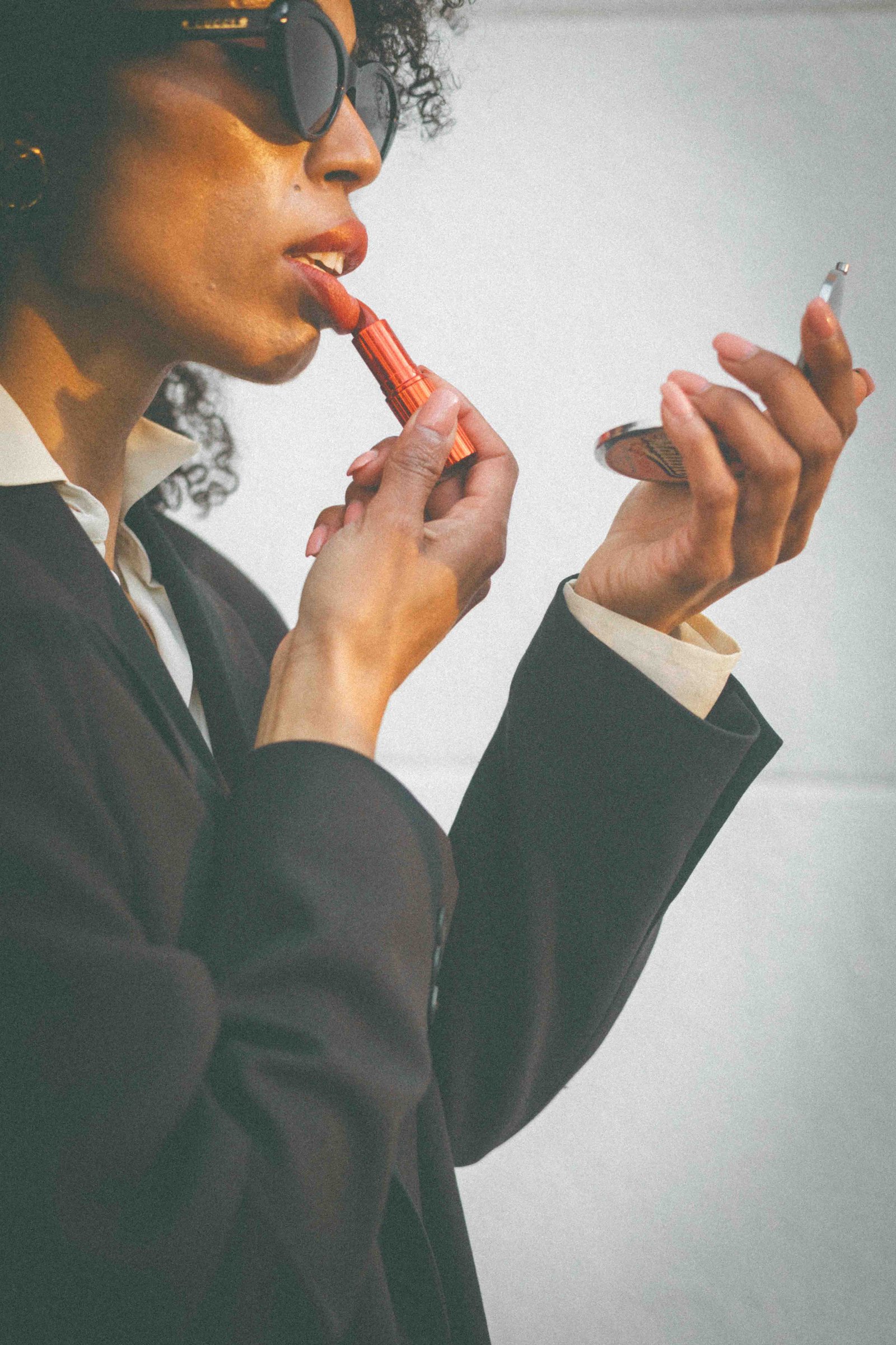 Black woman applying red lipstick in mirror wearing black blazer, intimate detail shot, dressing with intention