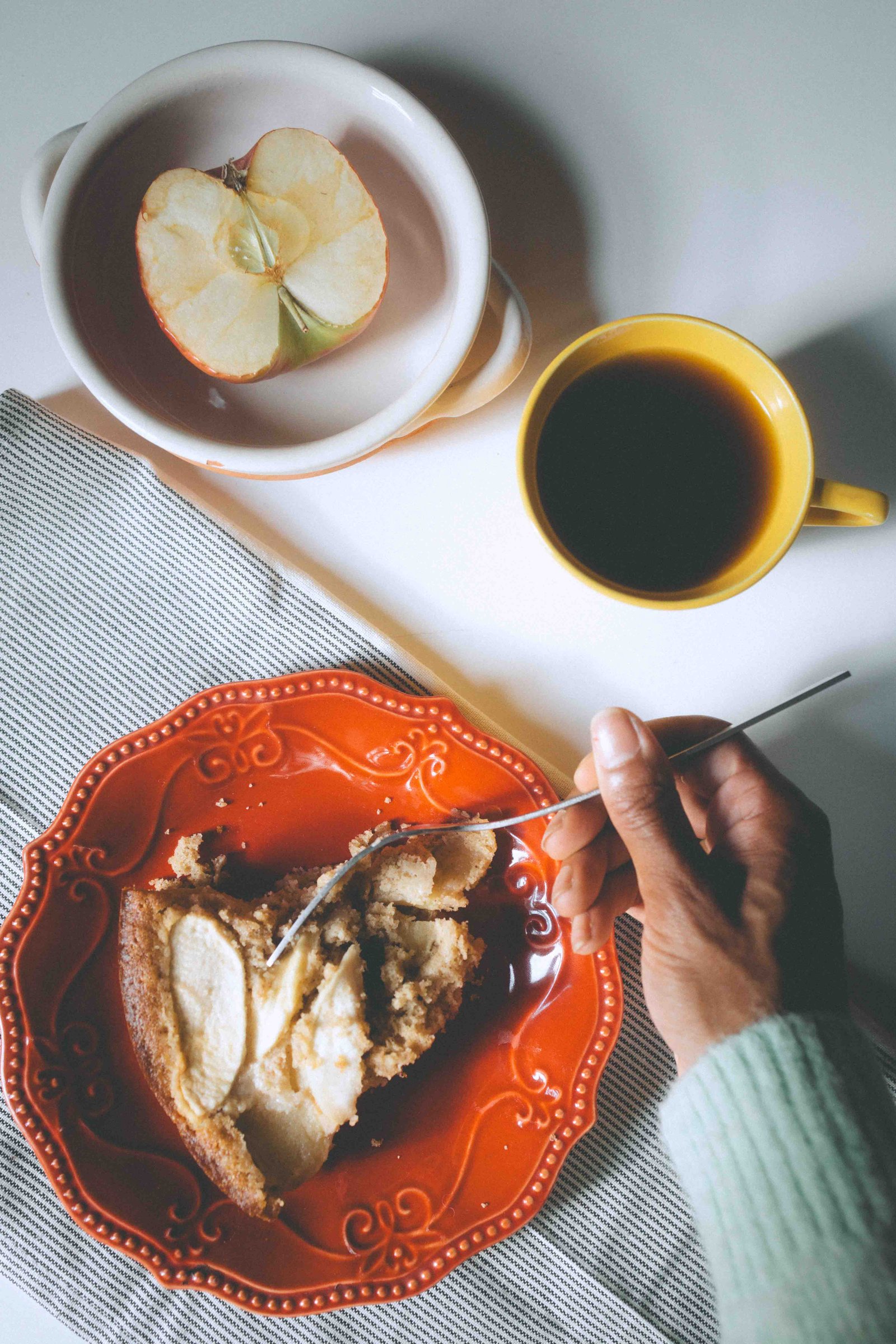 Hand holding a fork over a slice of apple cake on an orange plate beside coffee and a halved apple.