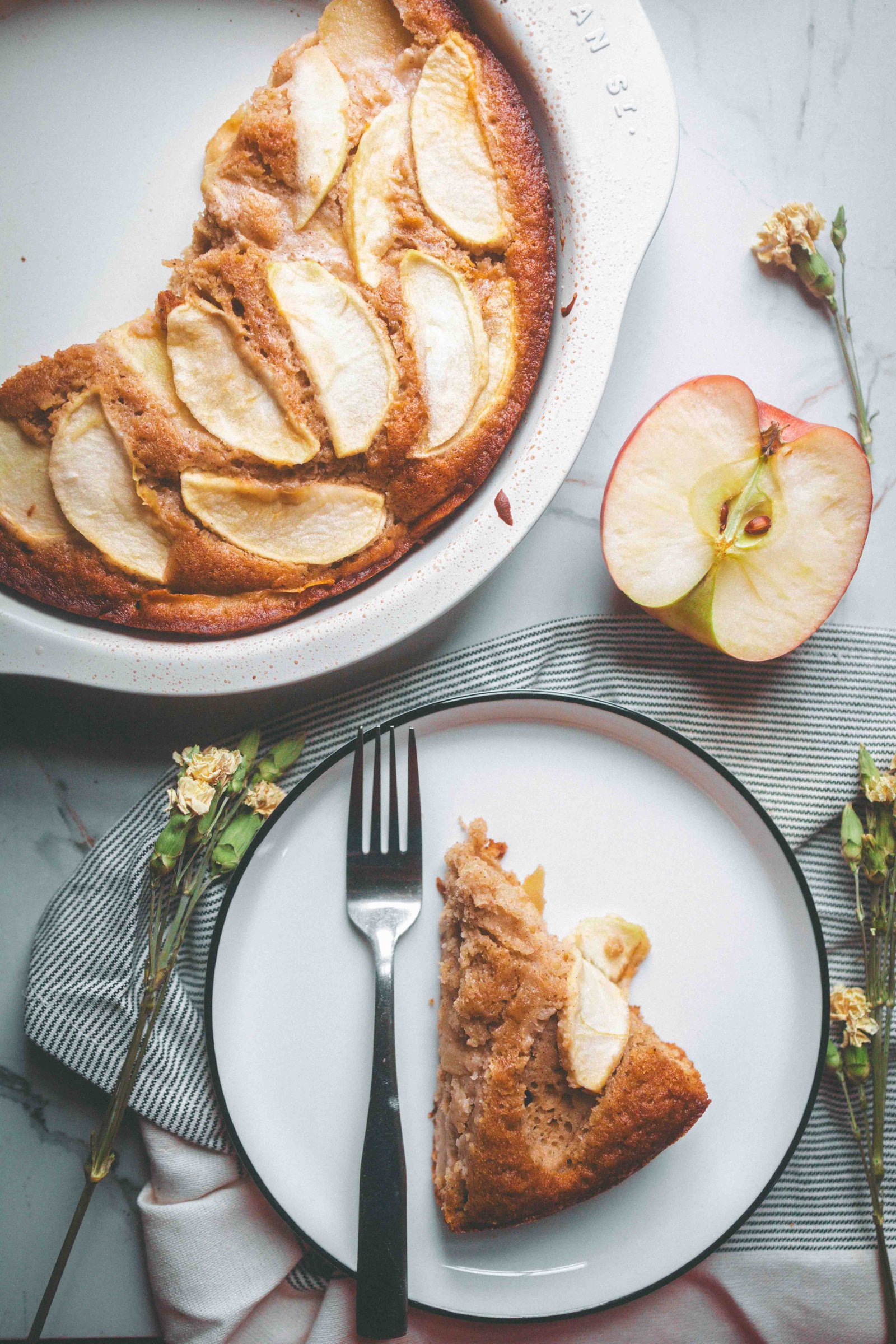 Brown butter apple cake in a baking dish with a plated slice, halved apple, fork, and dried flowers.