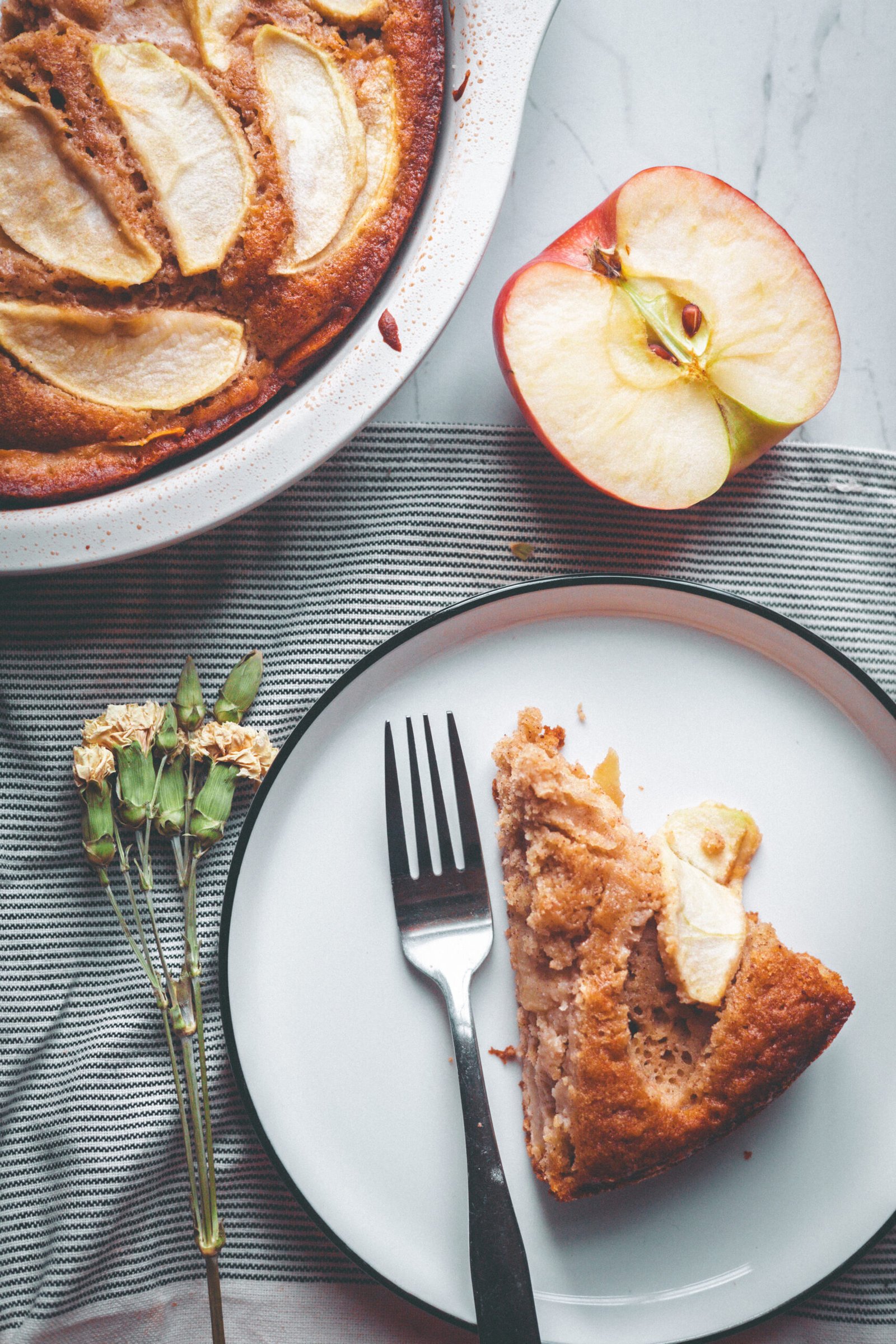Slice of brown butter apple cake on a white plate beside a halved red apple, dried flowers, and the remaining cake.