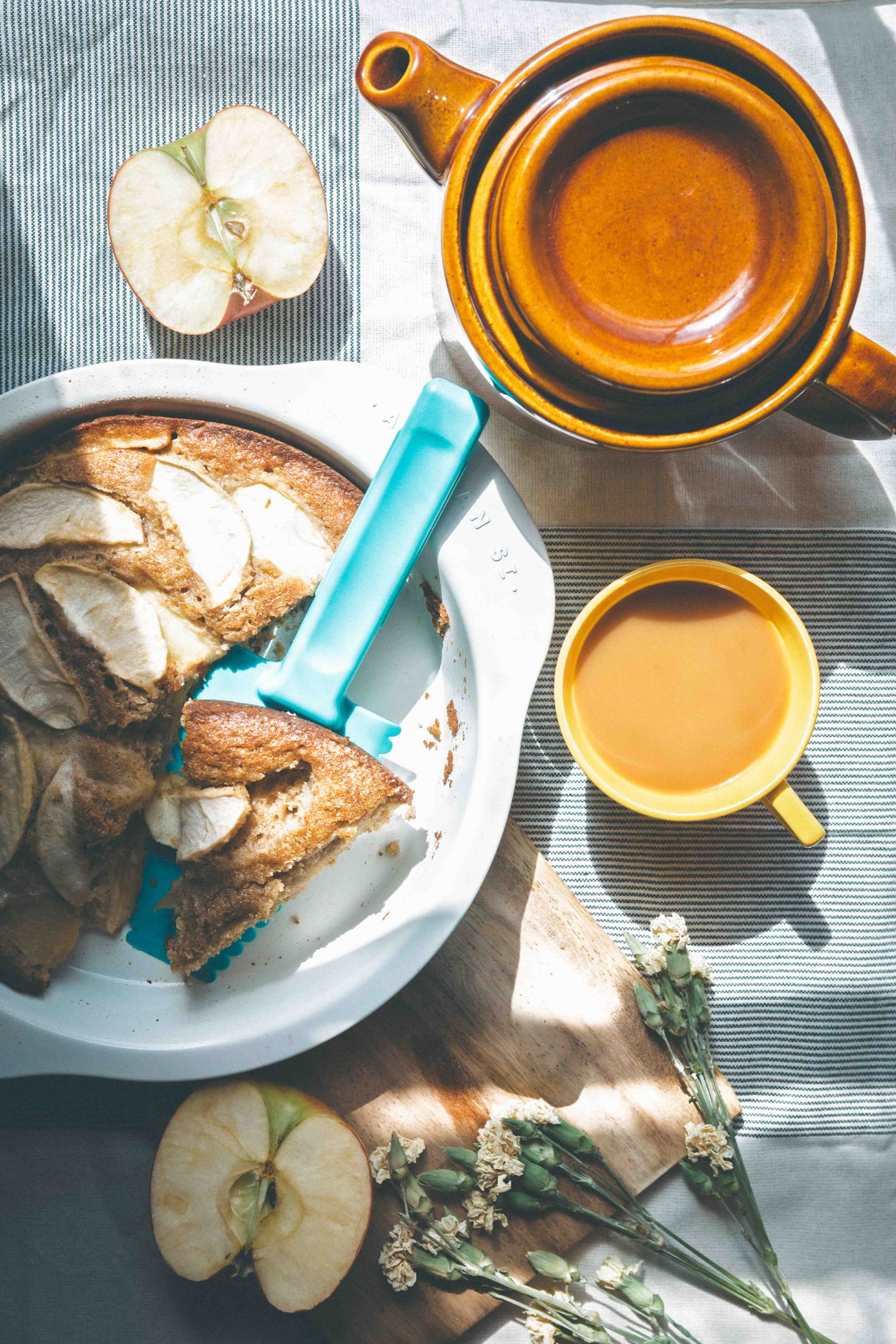 Brown butter apple cake served with coffee, a brown ceramic teapot, apples, and dried flowers in warm sunlight.