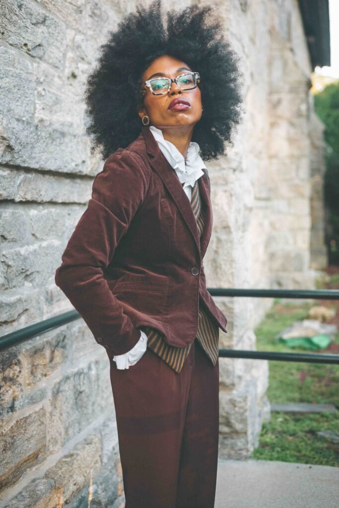 Editorial portrait of a woman in layered brown tailoring leaning beside a stone wall.