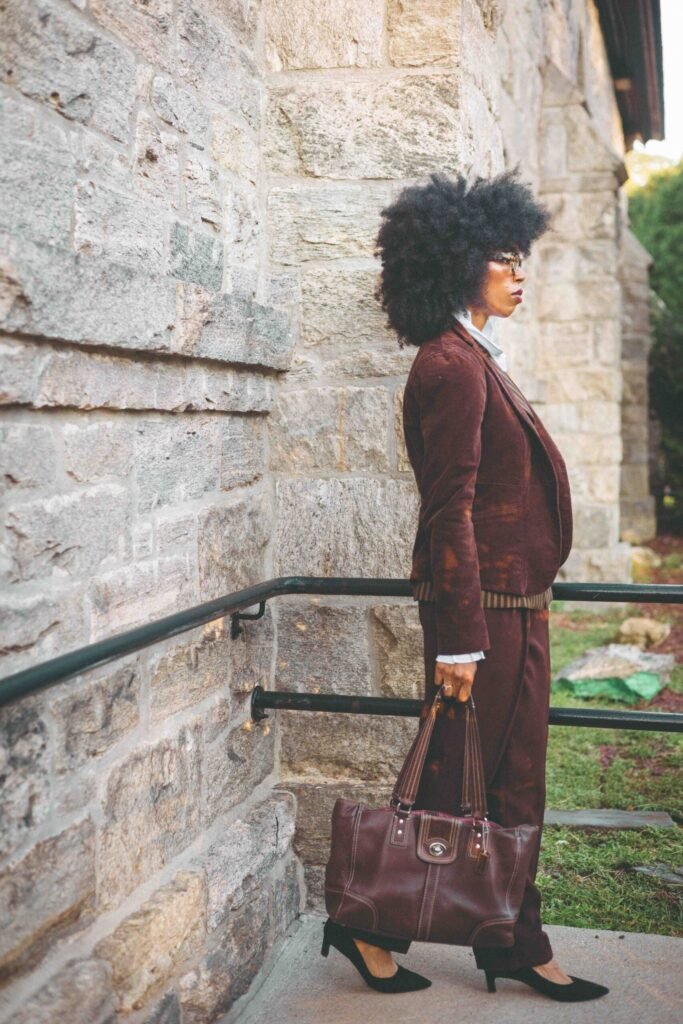 Side-profile portrait of a woman wearing brown corduroy tailoring beside a stone wall.