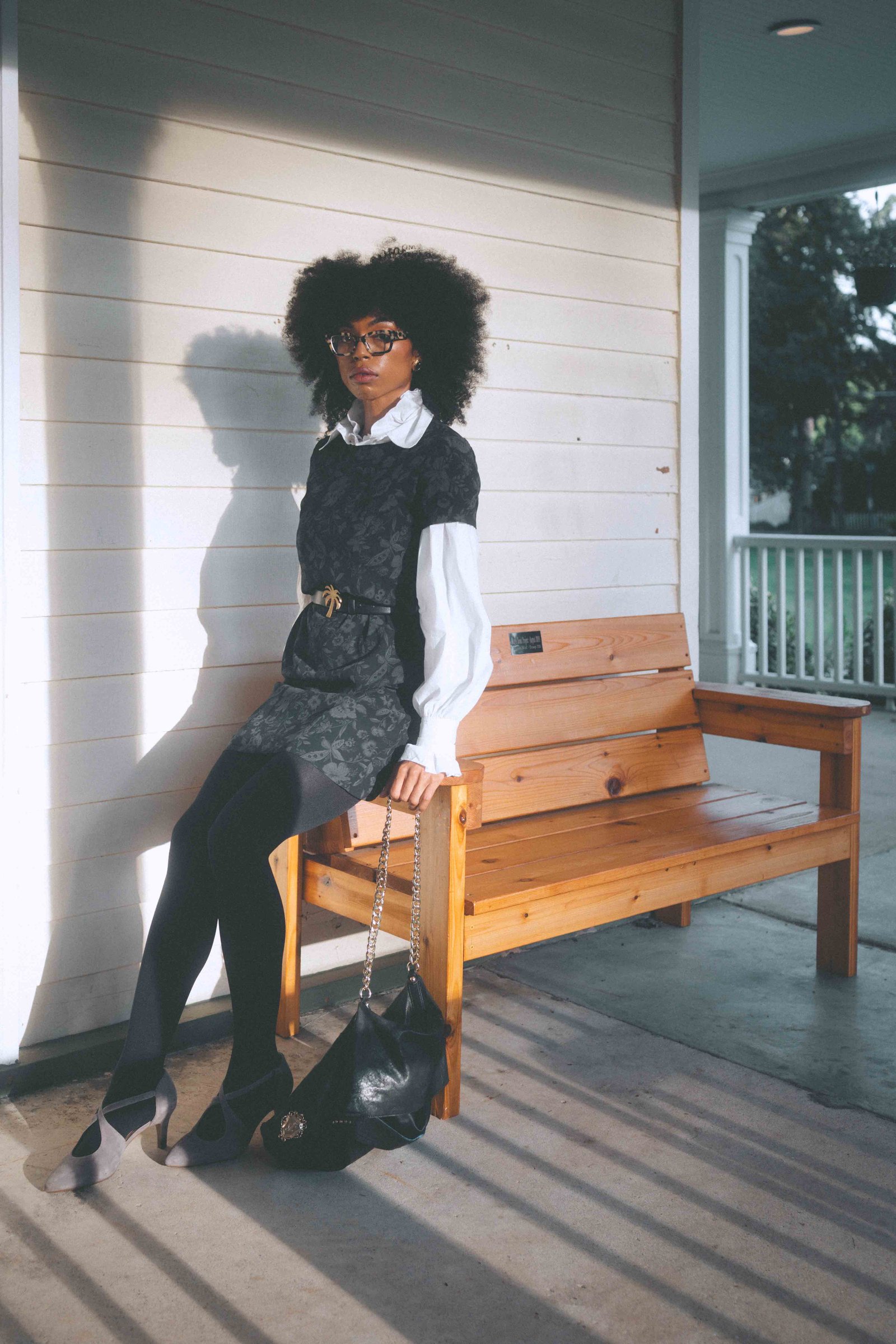 Woman seated on a porch bench wearing a belted dark floral dress over a white blouse