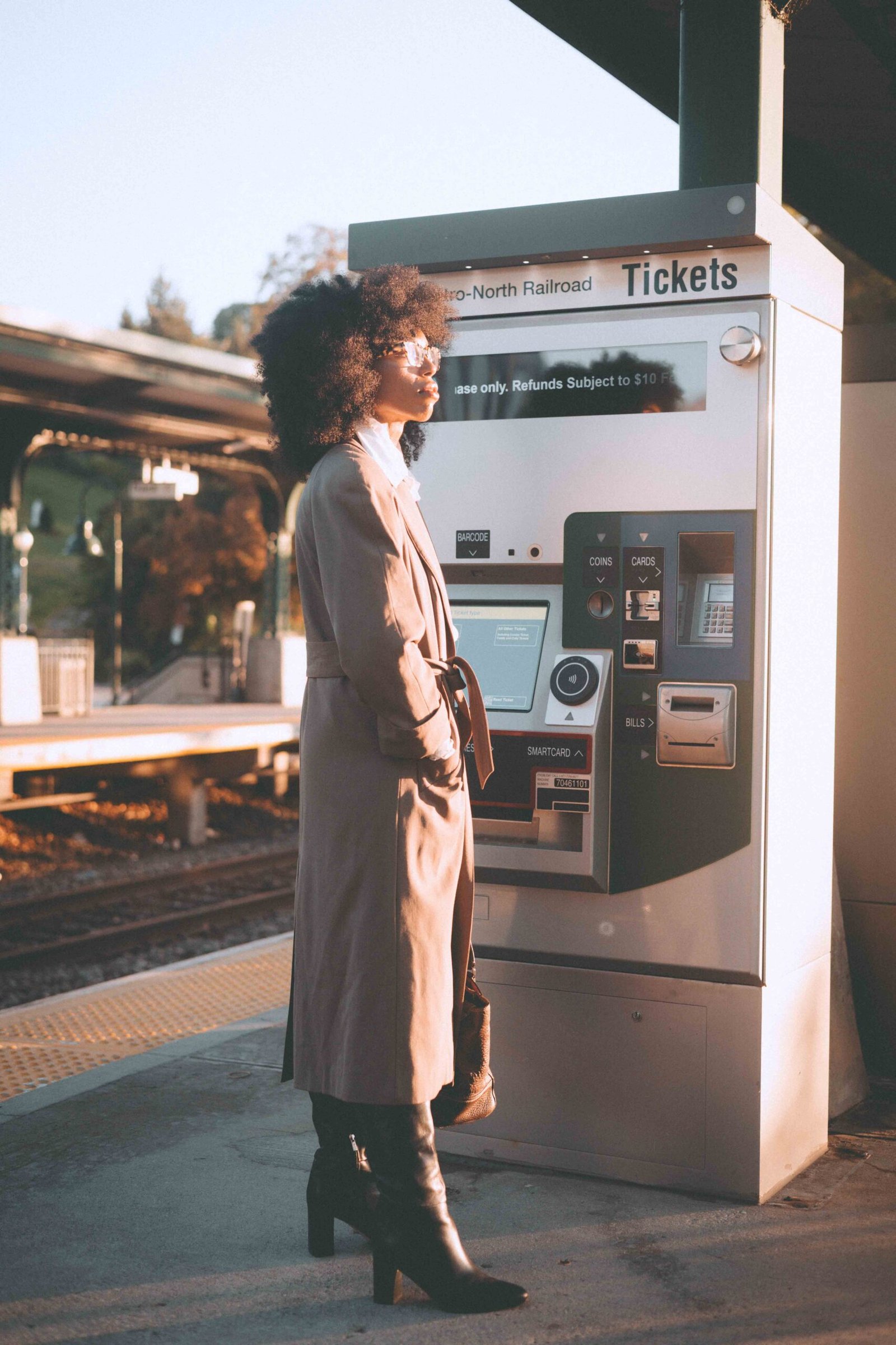 Side profile of a woman in a vintage trench coat standing beside a train-station ticket machine