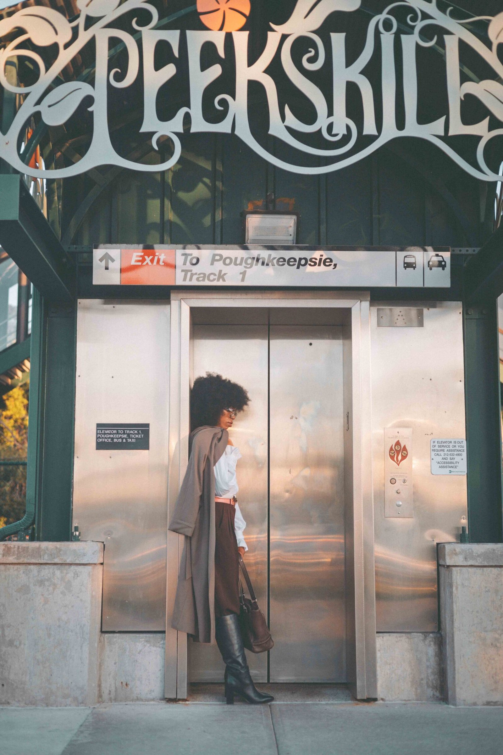 Full-length portrait of a woman in a vintage trench coat beneath the Peekskill station sign