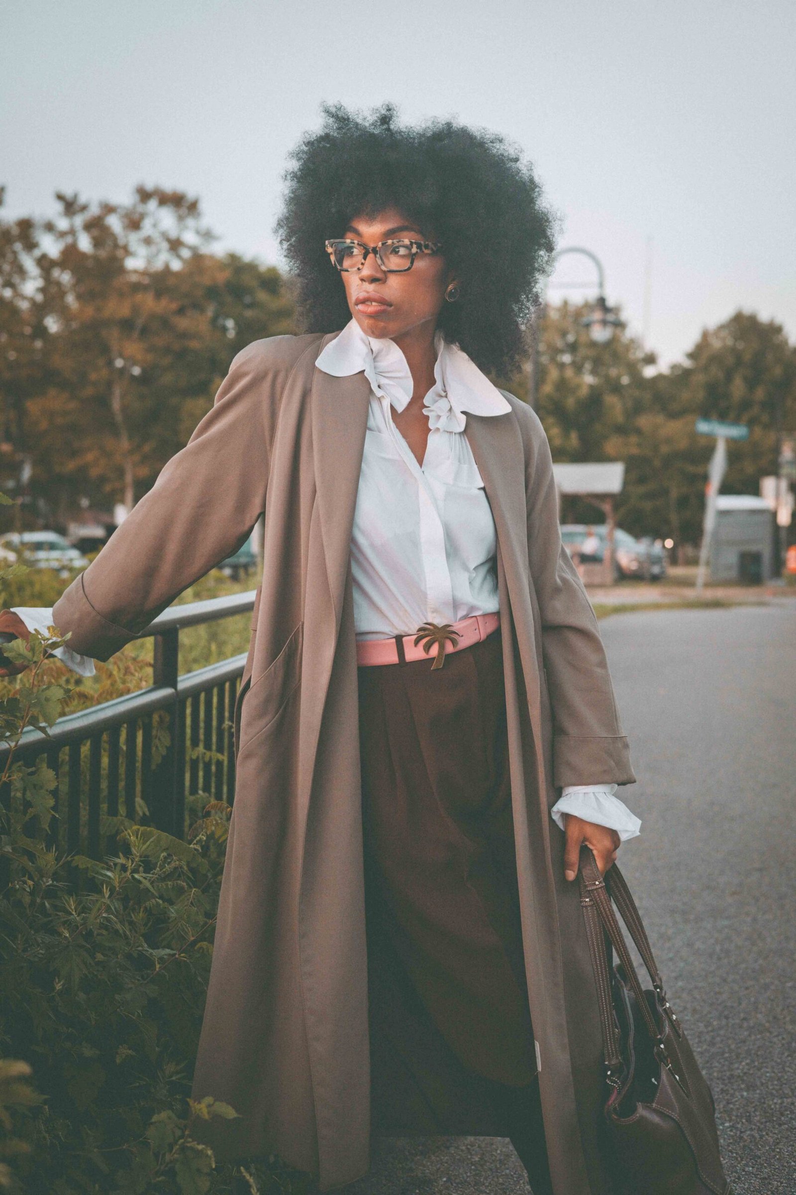 Woman wearing a taupe trench coat, white poet blouse and chocolate trousers near a train station