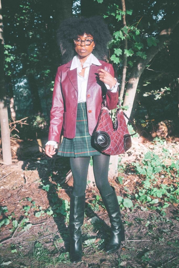 Woman standing in a wooded setting wearing a burgundy leather blazer and plaid wool skirt